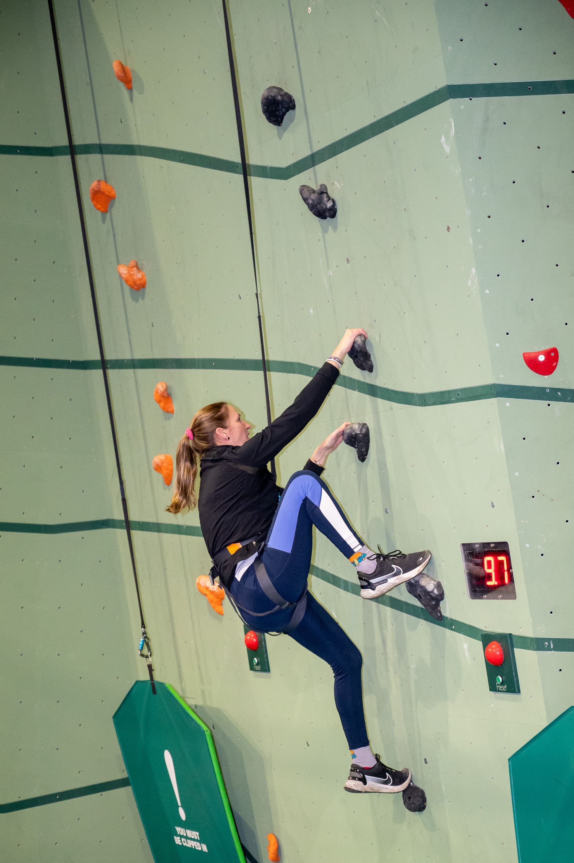 Woman climbing an indoor rock wall, wearing harness and athletic wear.