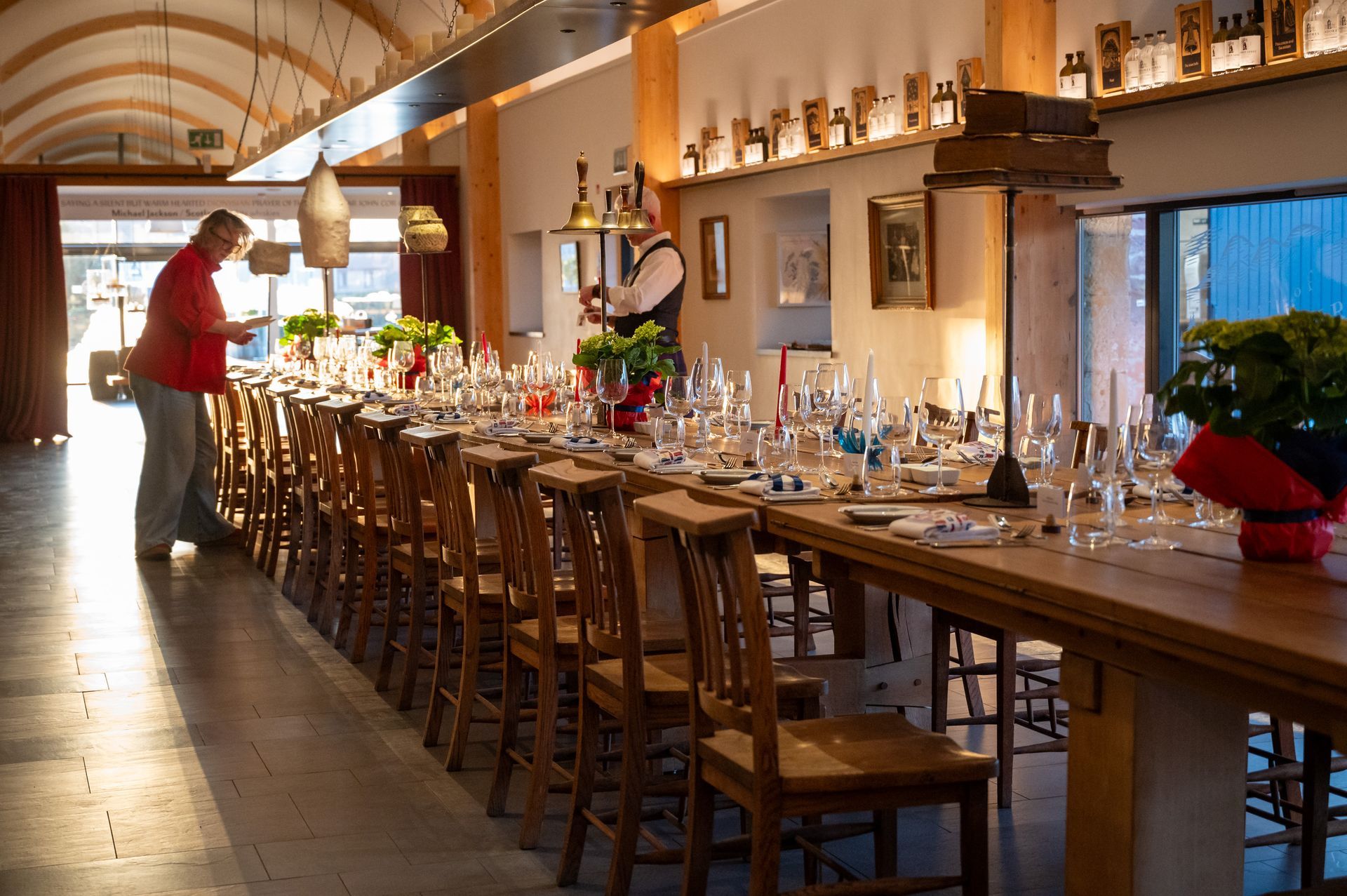 Long wooden table set for a meal, rows of chairs, two people at each end.