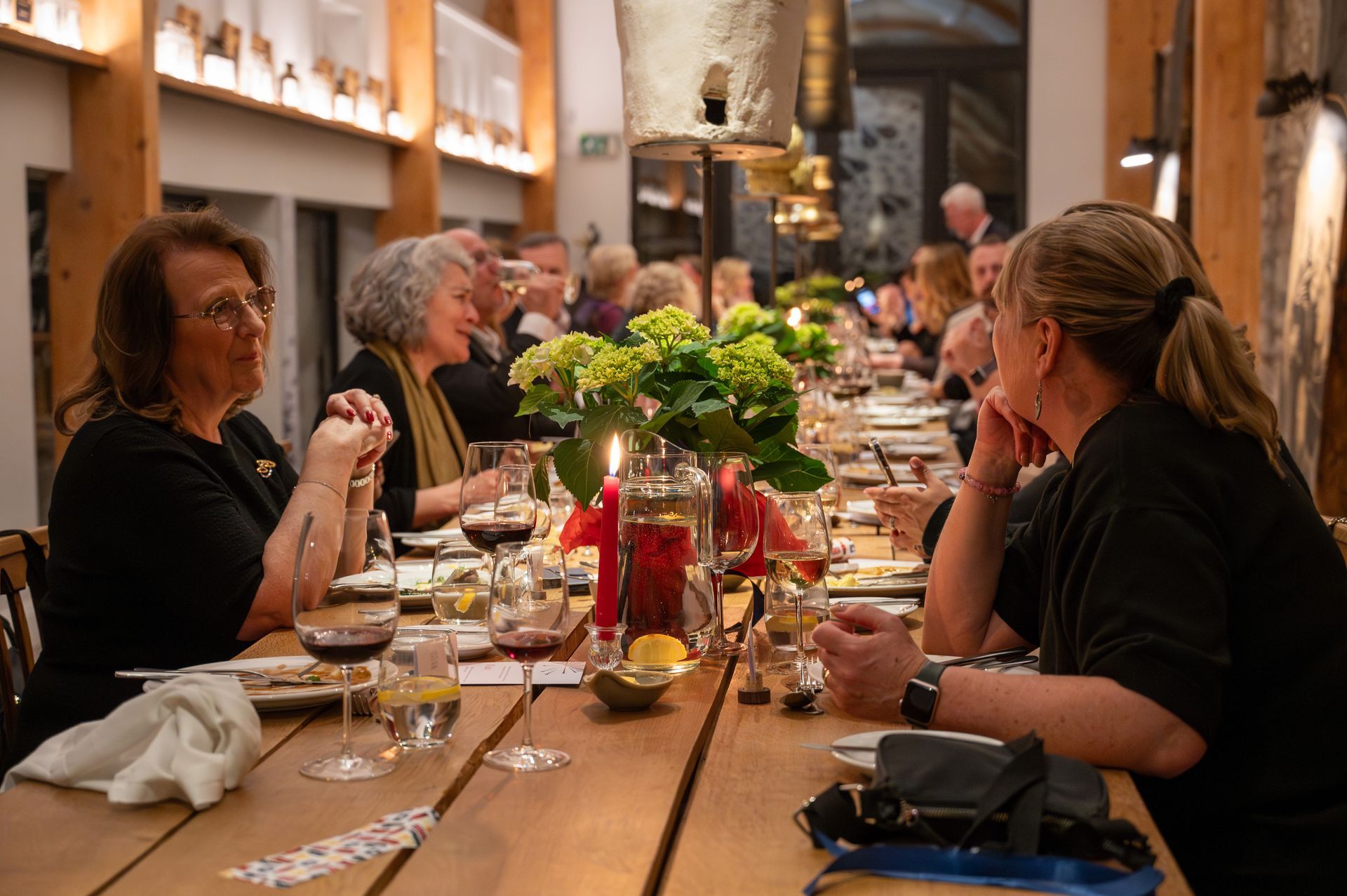 People seated at a long wooden table, enjoying a meal. Candles, flowers and wine glasses are present.