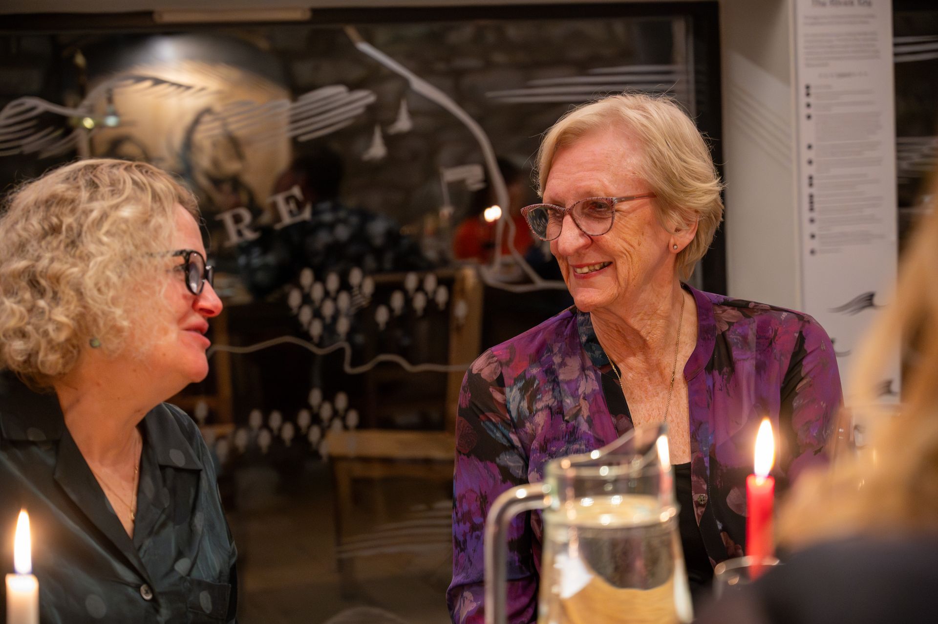 Two women talking at a candlelit table, smiling. One wears glasses, a purple shirt. Behind them, a decorated window.