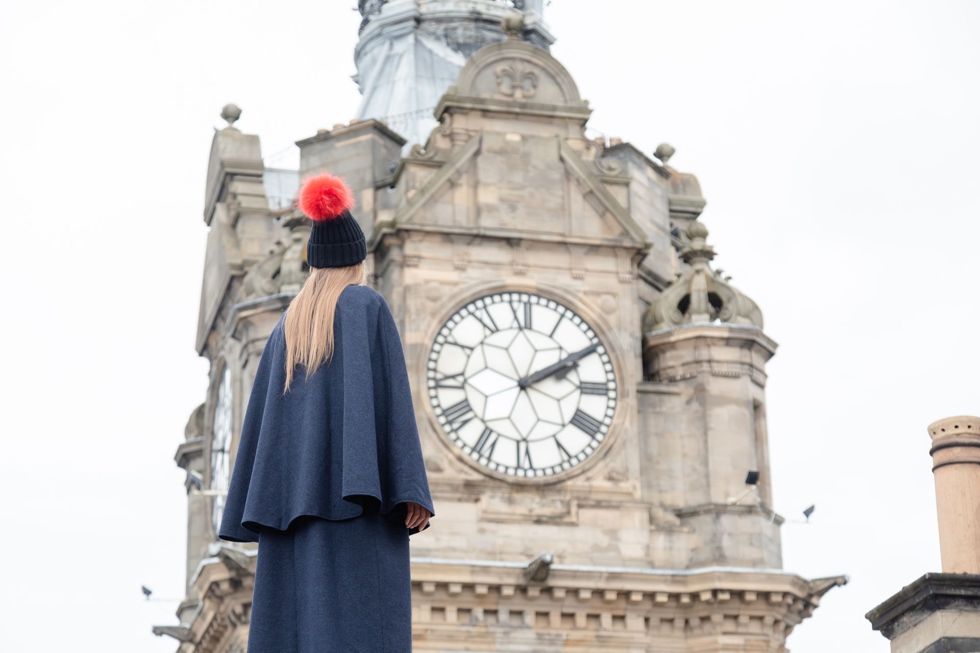 Woman in blue cape and hat with pom-pom facing a large clock tower.