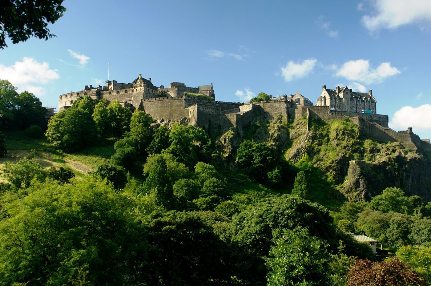 Edinburgh Castle atop a green, rocky hill, under a blue sky with clouds.