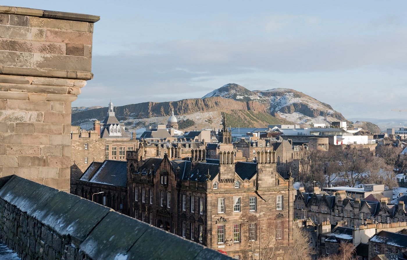 Edinburgh cityscape with castle walls in the foreground, Arthur's Seat in the distance, snowy rooftops.