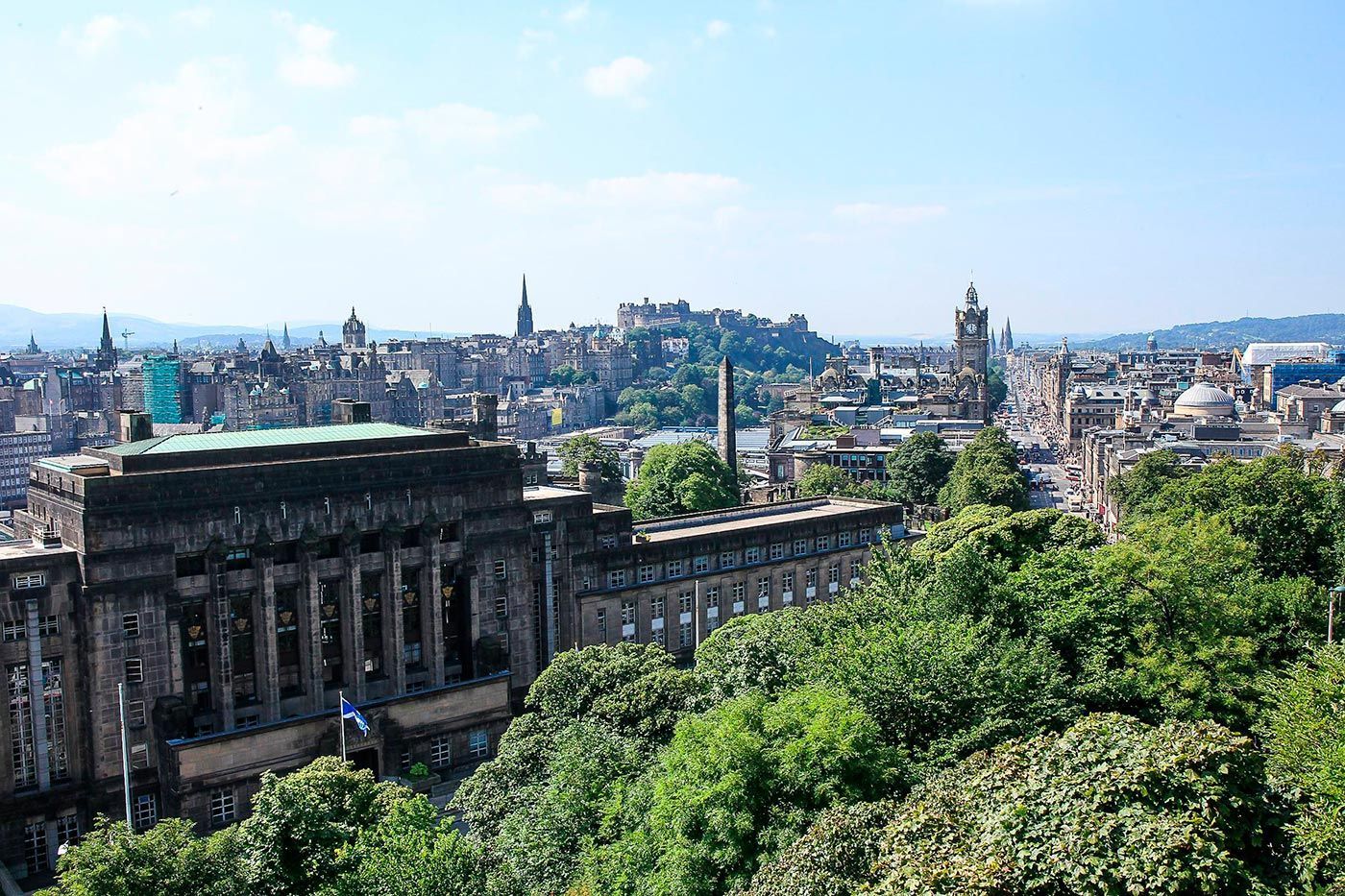 Edinburgh cityscape with castle on a hill, buildings, and green trees under a blue sky.