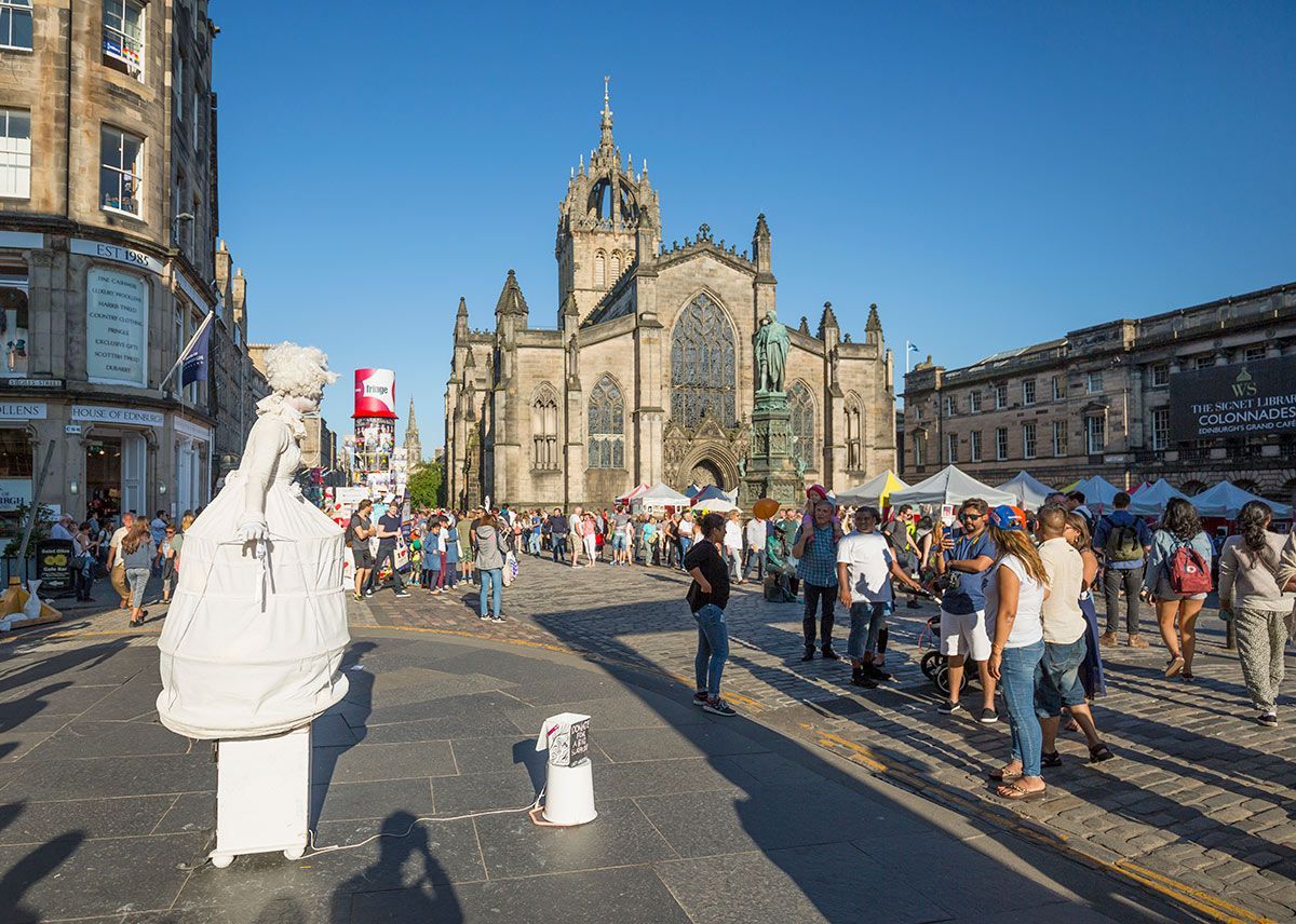Crowd in Edinburgh's Royal Mile, with St. Giles' Cathedral in the background, a street performer, and market stalls.