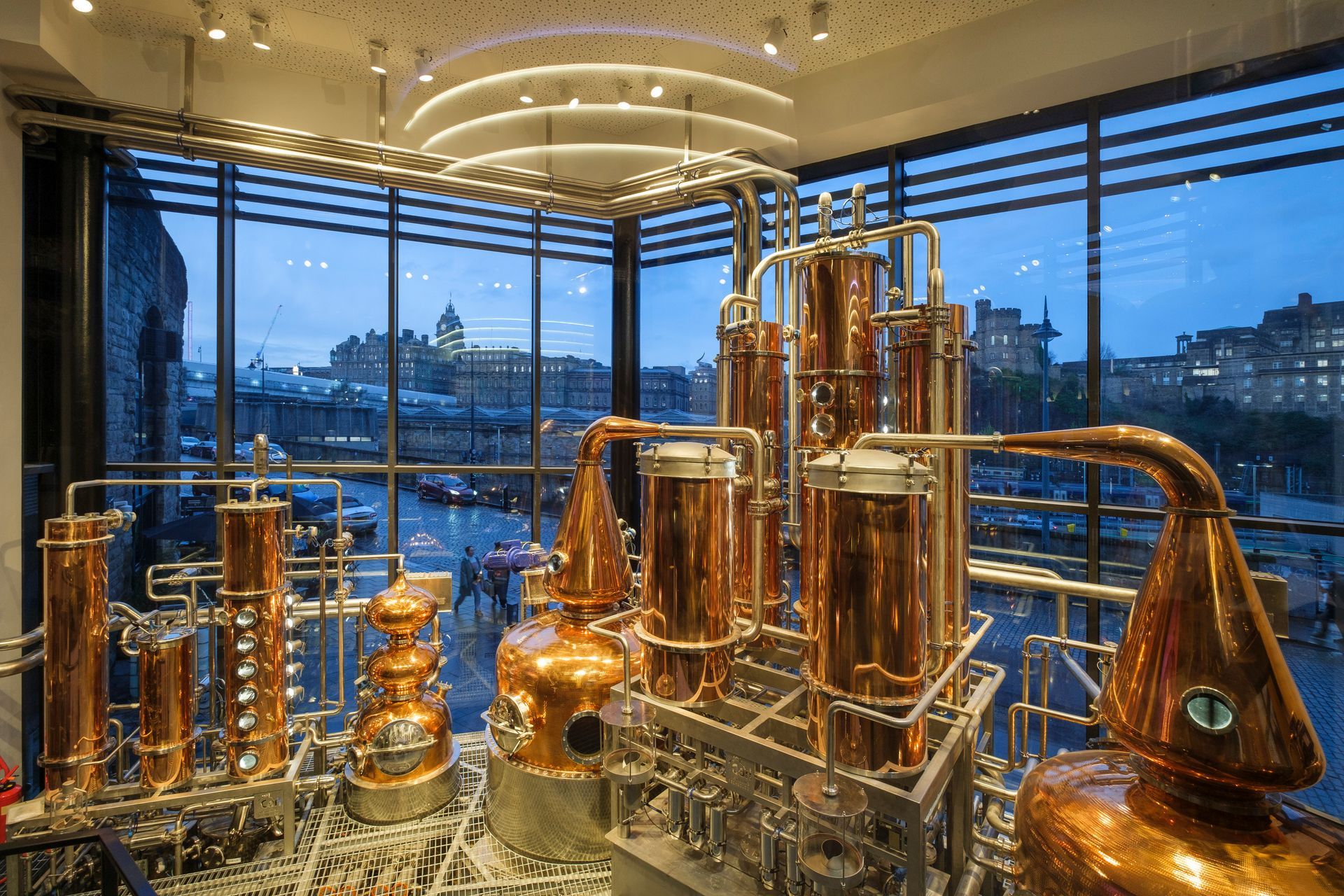 Copper stills in a distillery, viewed through large windows with a city backdrop.