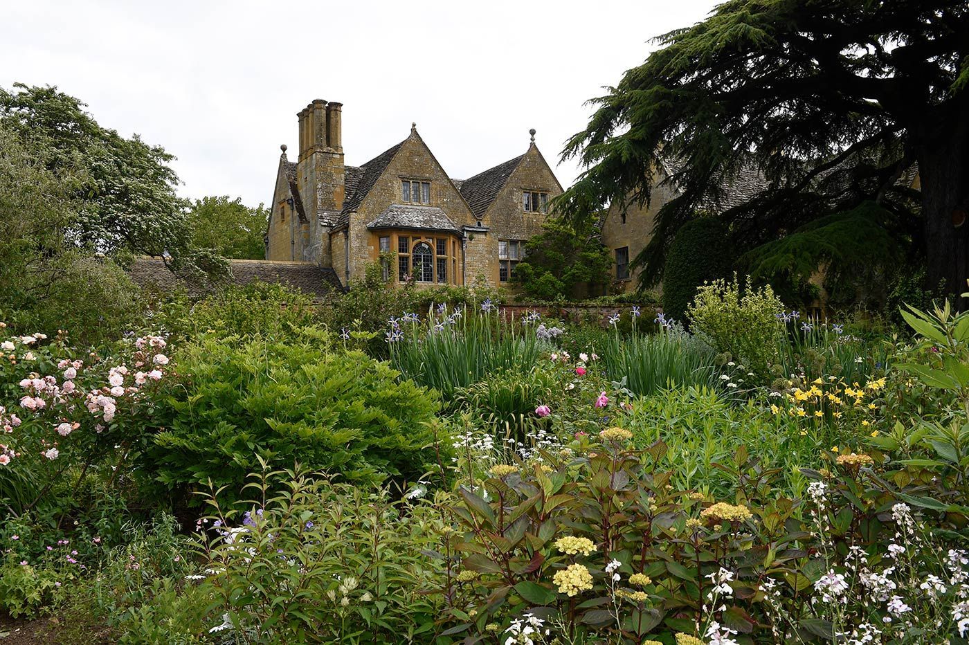 Stone manor house behind a vibrant flower garden with lush greenery and a cloudy sky.
