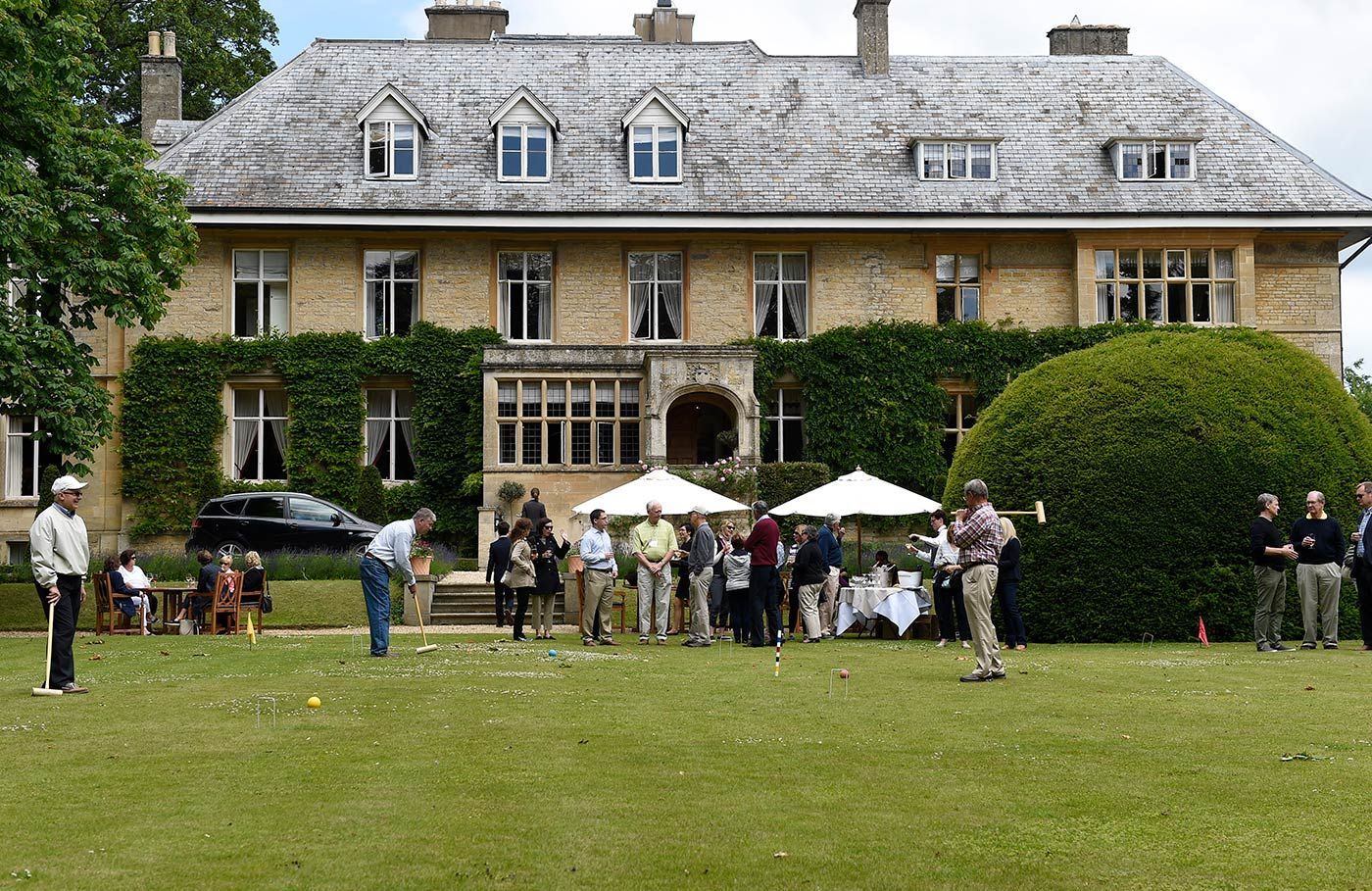 Large house with people playing croquet on lawn. Guests stand near tables.