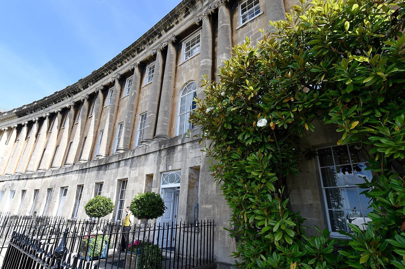 Curved row of light-colored buildings with arched windows, partially obscured by green foliage, under a blue sky.