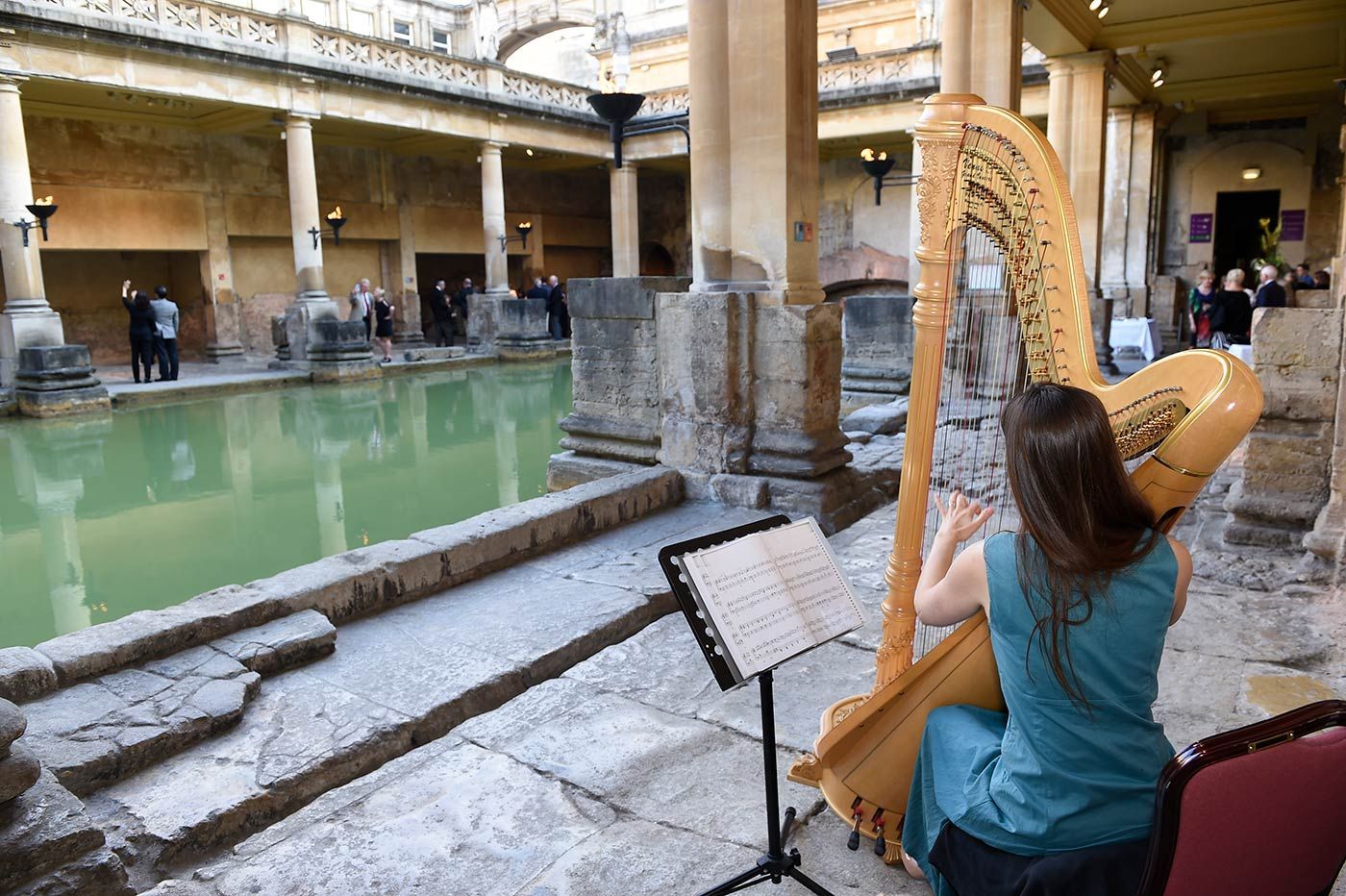Woman plays a harp in an ancient Roman bathhouse. The pool has greenish water, stone pillars.