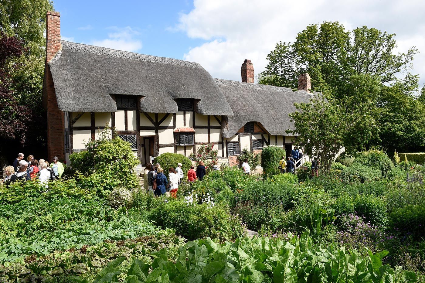 Thatched-roof cottage with garden and people gathered in front.
