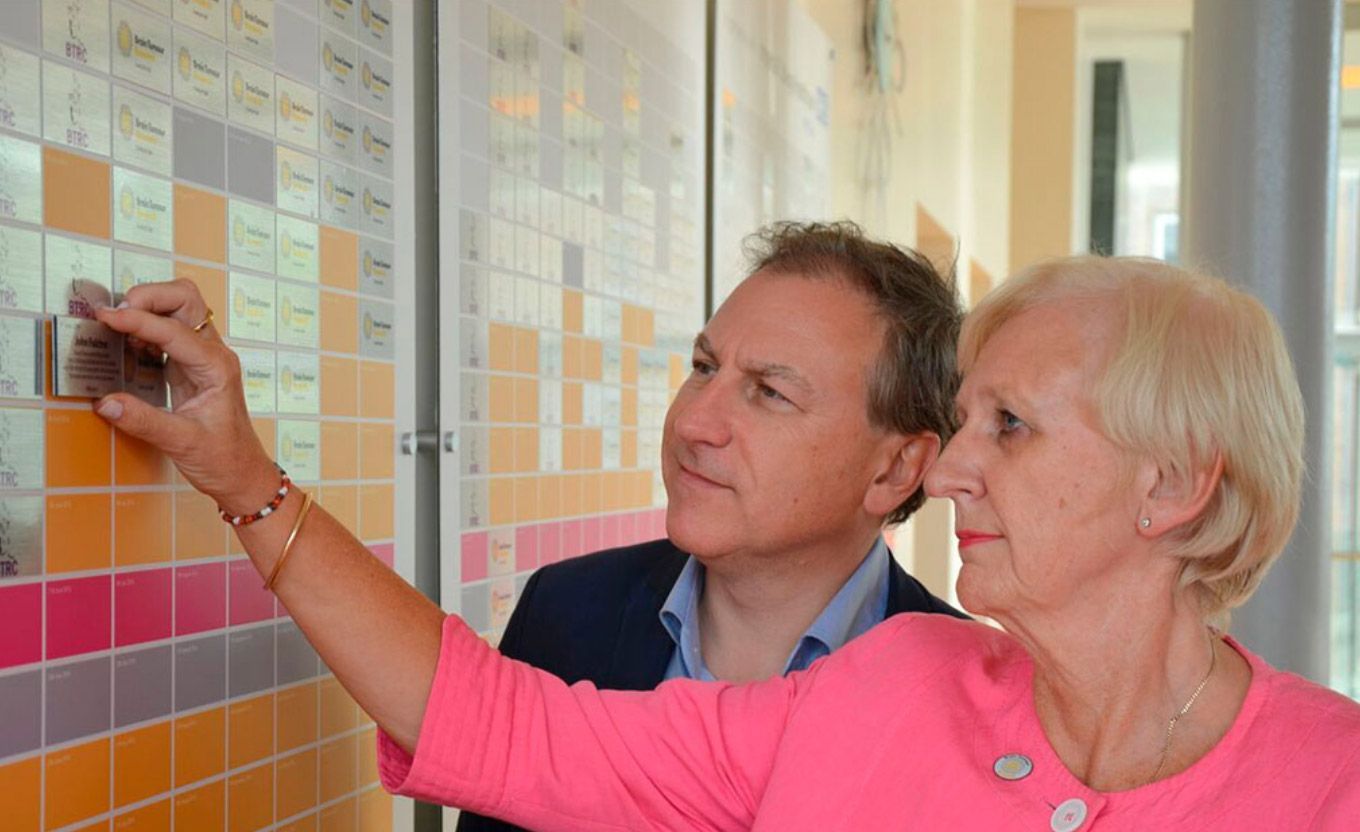 Two people looking at a wall of square tiles, one pointing. Light-filled hallway.