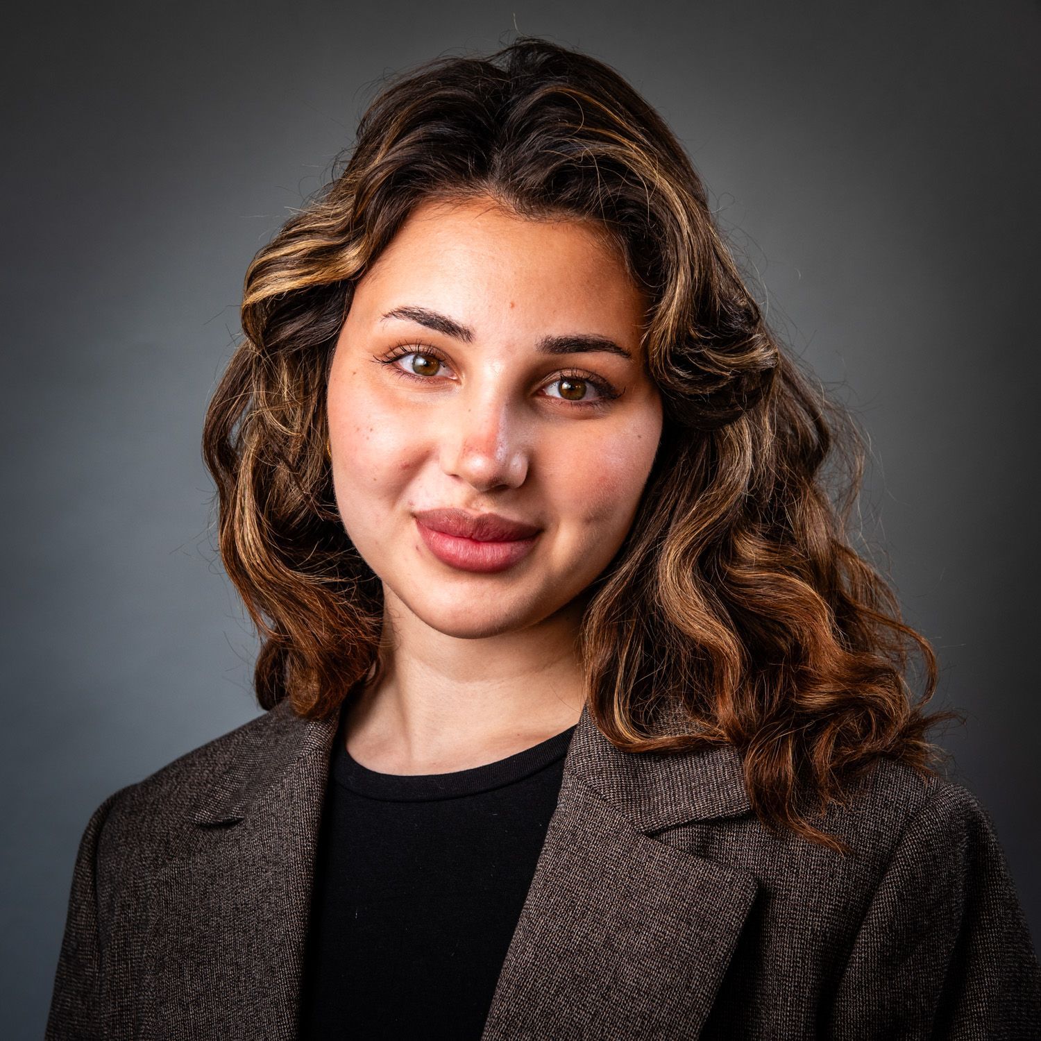 Woman with curly brown hair, wearing a black top and brown blazer, smiling, studio shot.