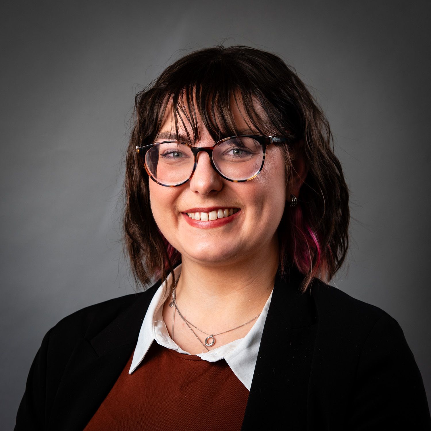 Woman with glasses smiles, wearing a blazer over a white collared shirt and rust-colored top, against a gray backdrop.