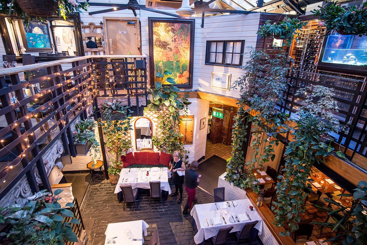 Courtyard restaurant with tables, greenery, and hanging lights. People stand near tables, a painting on the wall.