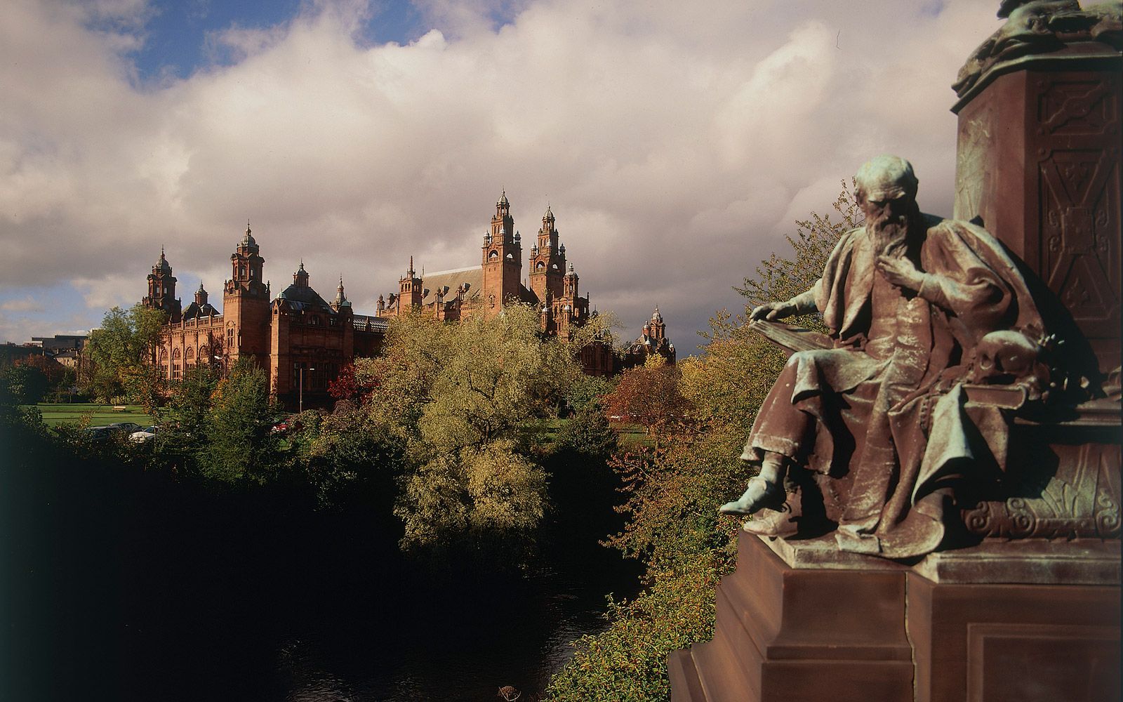Statue of a seated man, with a large ornate building in the background under a cloudy sky.