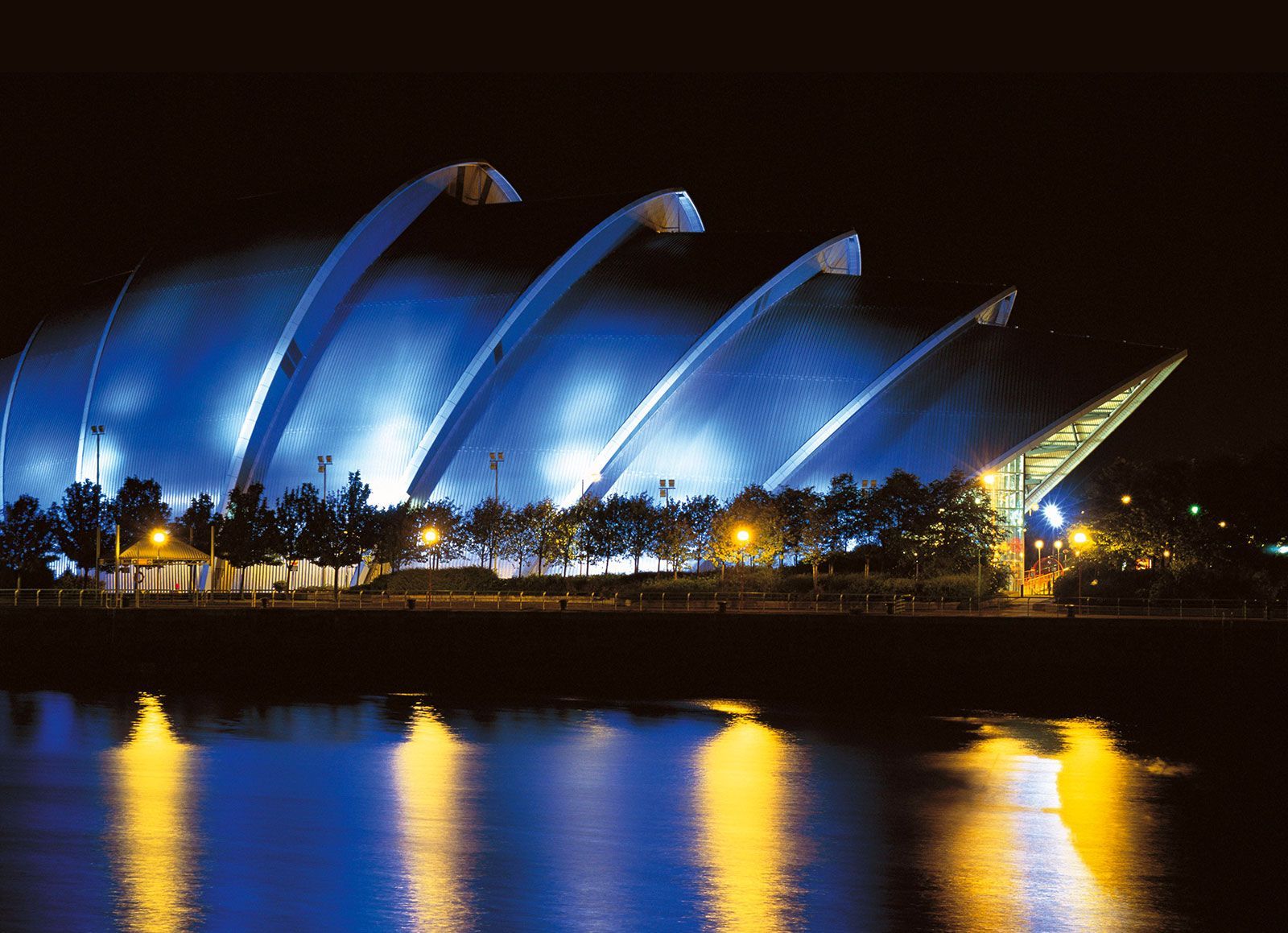 Blue-lit, arched roof structure at night reflects in dark water; trees line the foreground.