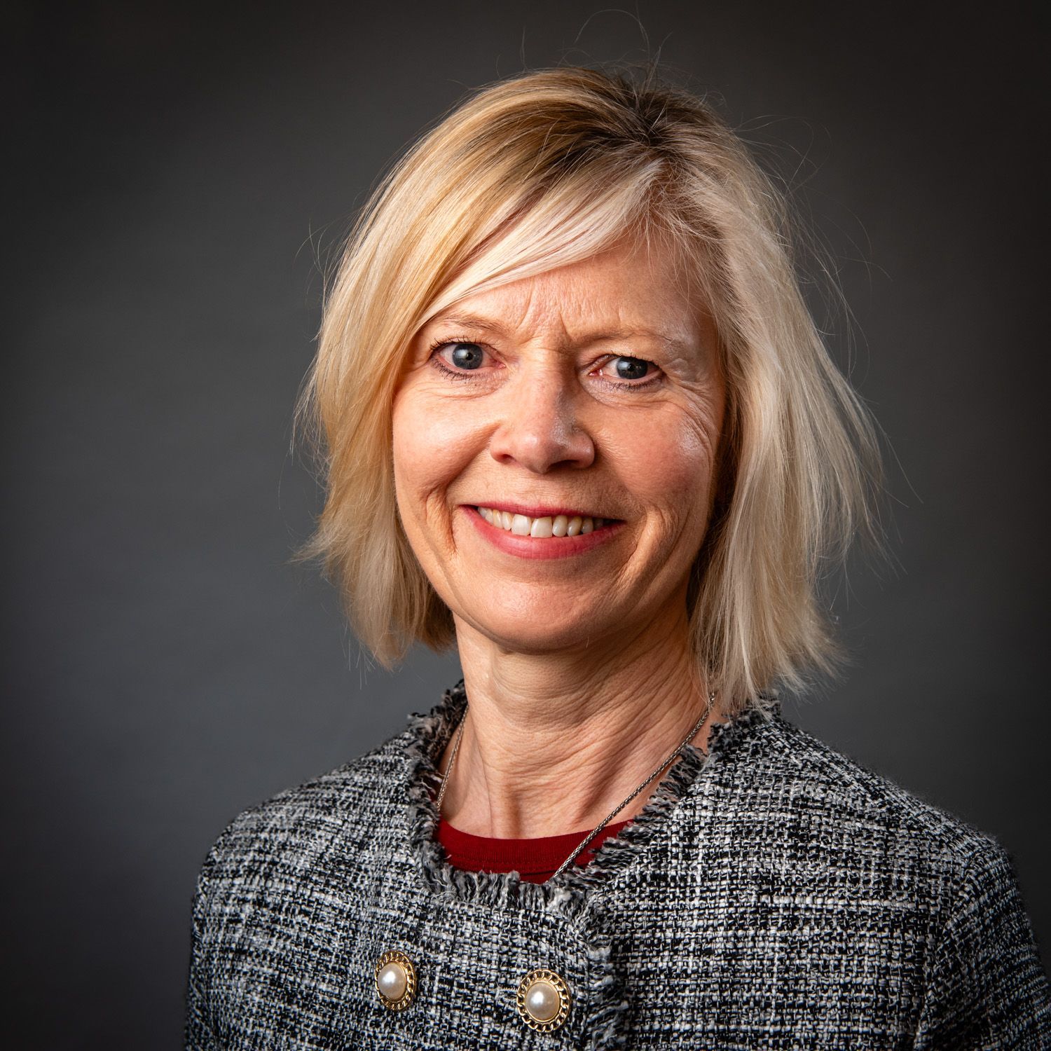 Woman with blonde hair wearing a patterned blazer smiling at the camera against a grey background.