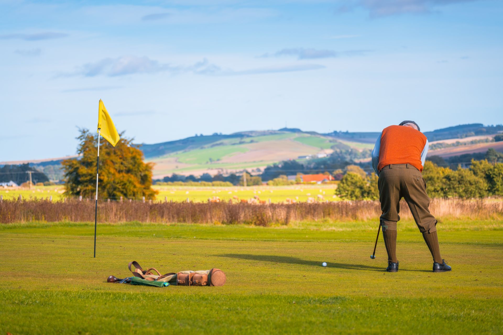 Golfer in orange vest swings club toward green with yellow flag; golf bag on the grass; rolling hills in background.