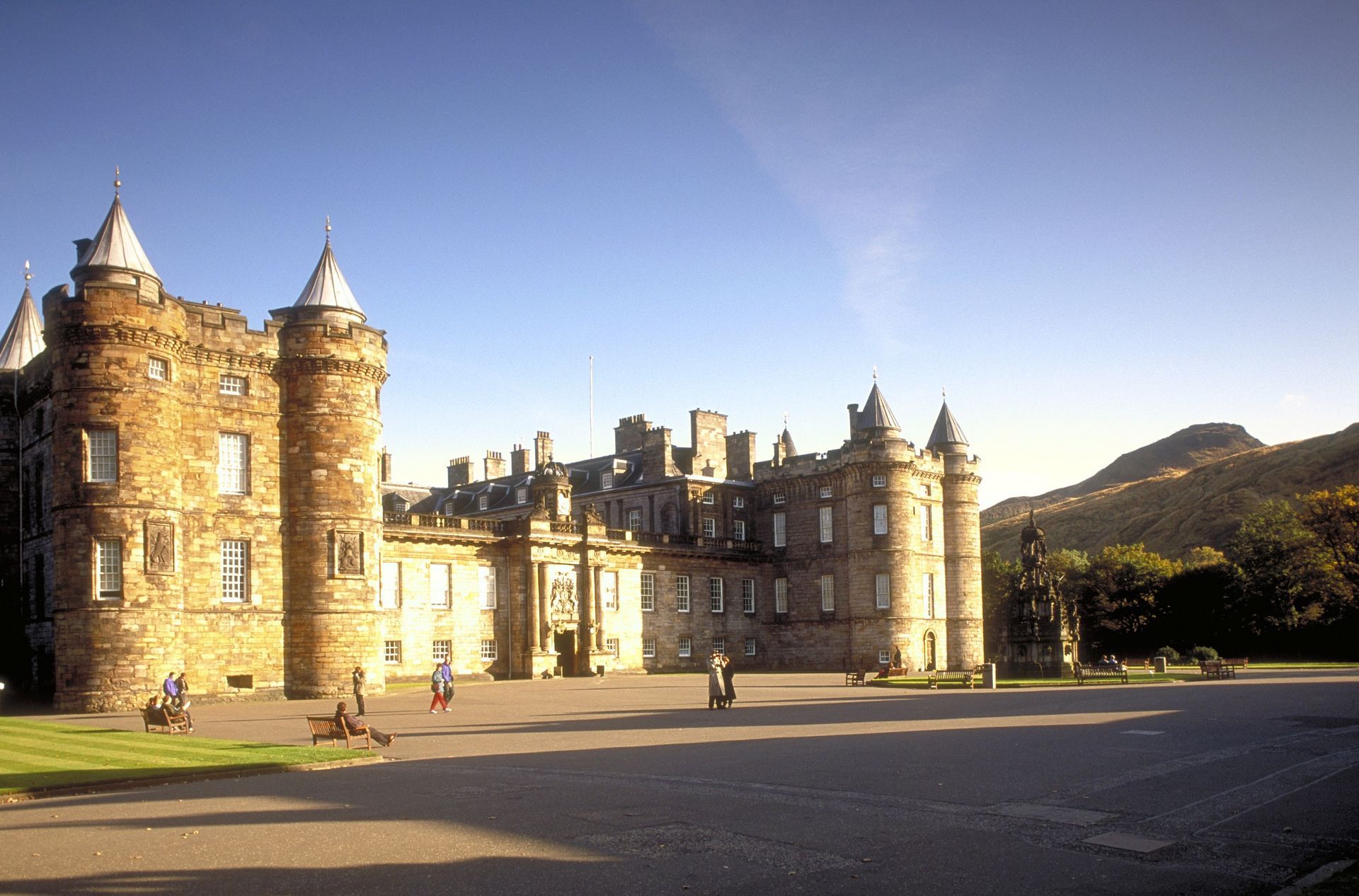 Palace of Holyroodhouse on a sunny day. Stone building with towers, courtyard, and figures walking.