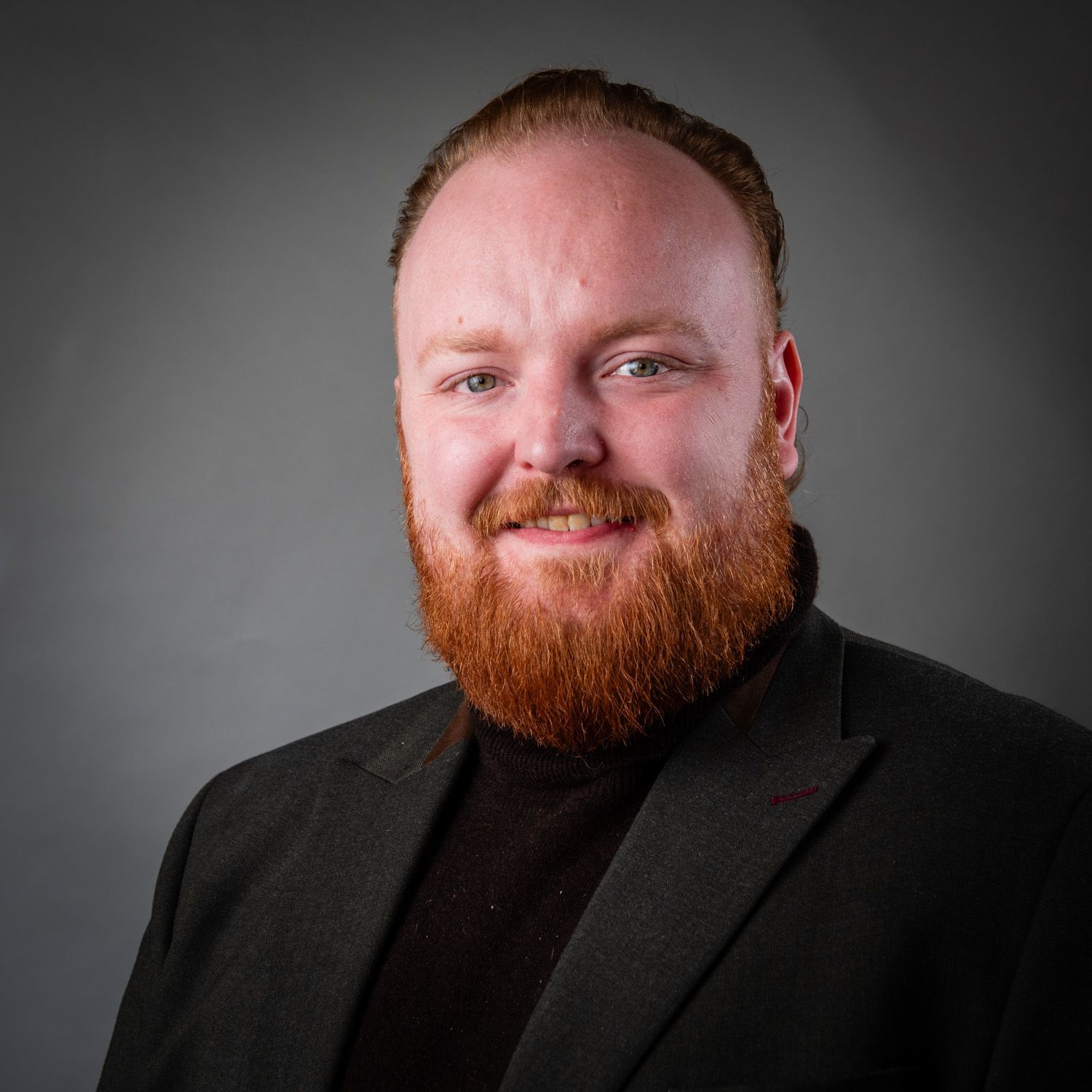 Man with red beard, wearing a black turtleneck and blazer, smiles against gray backdrop.
