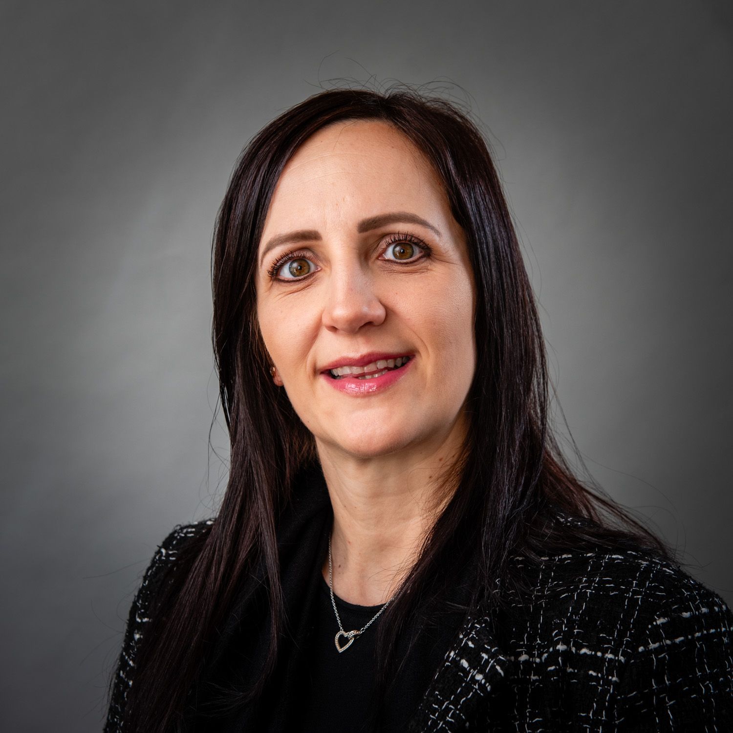 Woman with long dark hair, wearing a black blazer, smiles at the camera against a gray background.