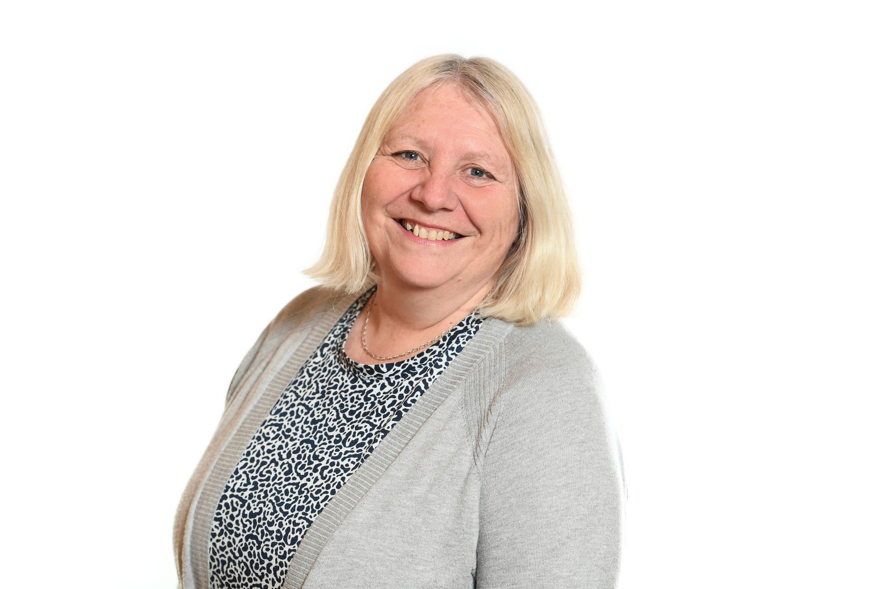 Woman with blonde hair, smiling, wearing a patterned shirt and gray cardigan, against a white background.
