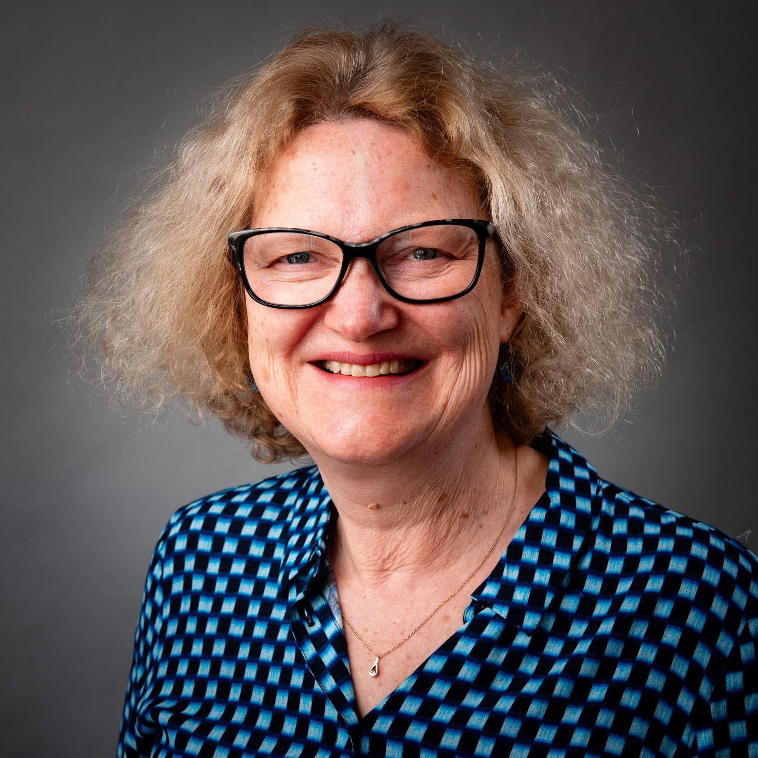 Woman with blonde hair and glasses smiles wearing a blue patterned shirt against a gray background.