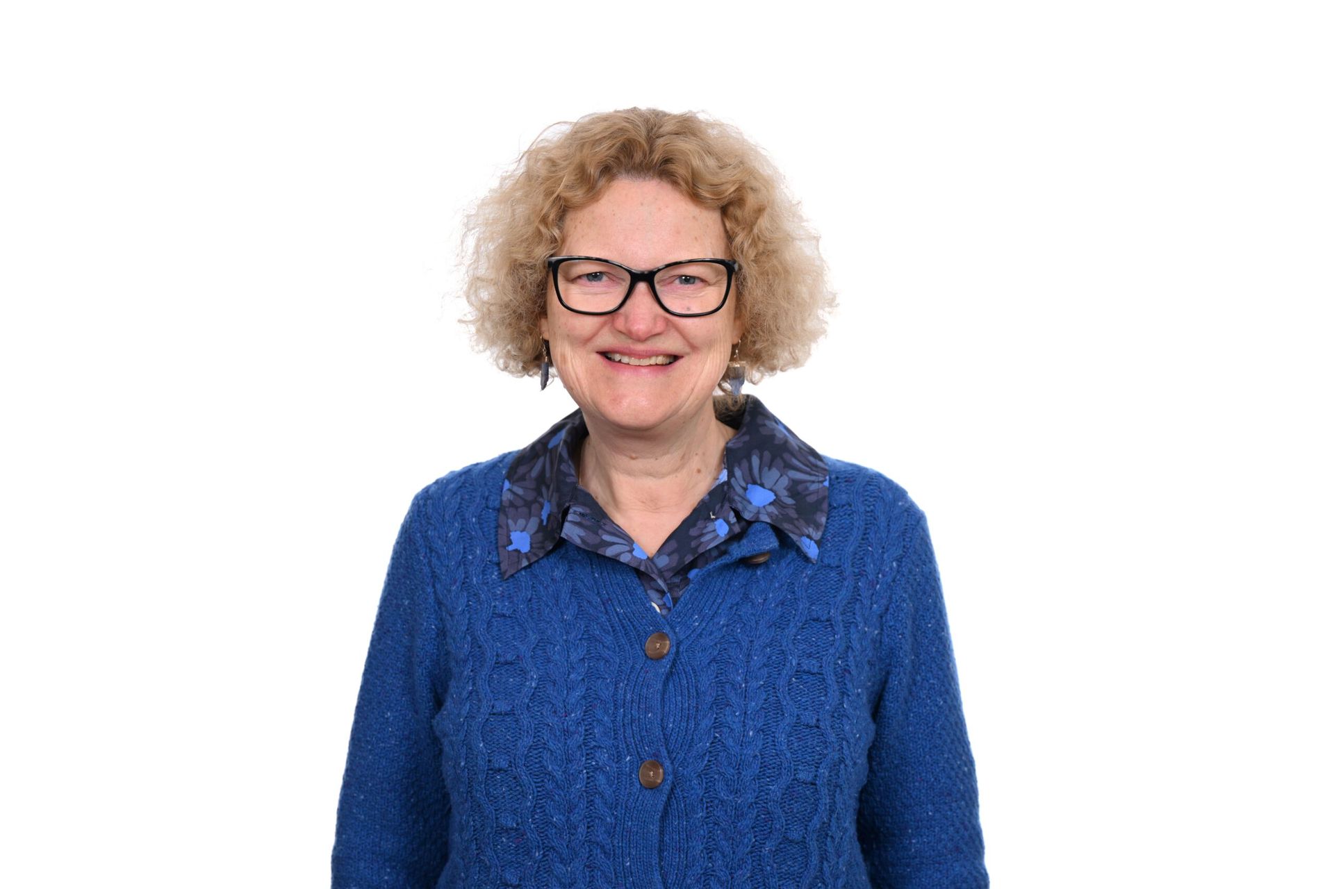 Woman with curly blonde hair and glasses wearing a blue sweater, smiling, against a white background.