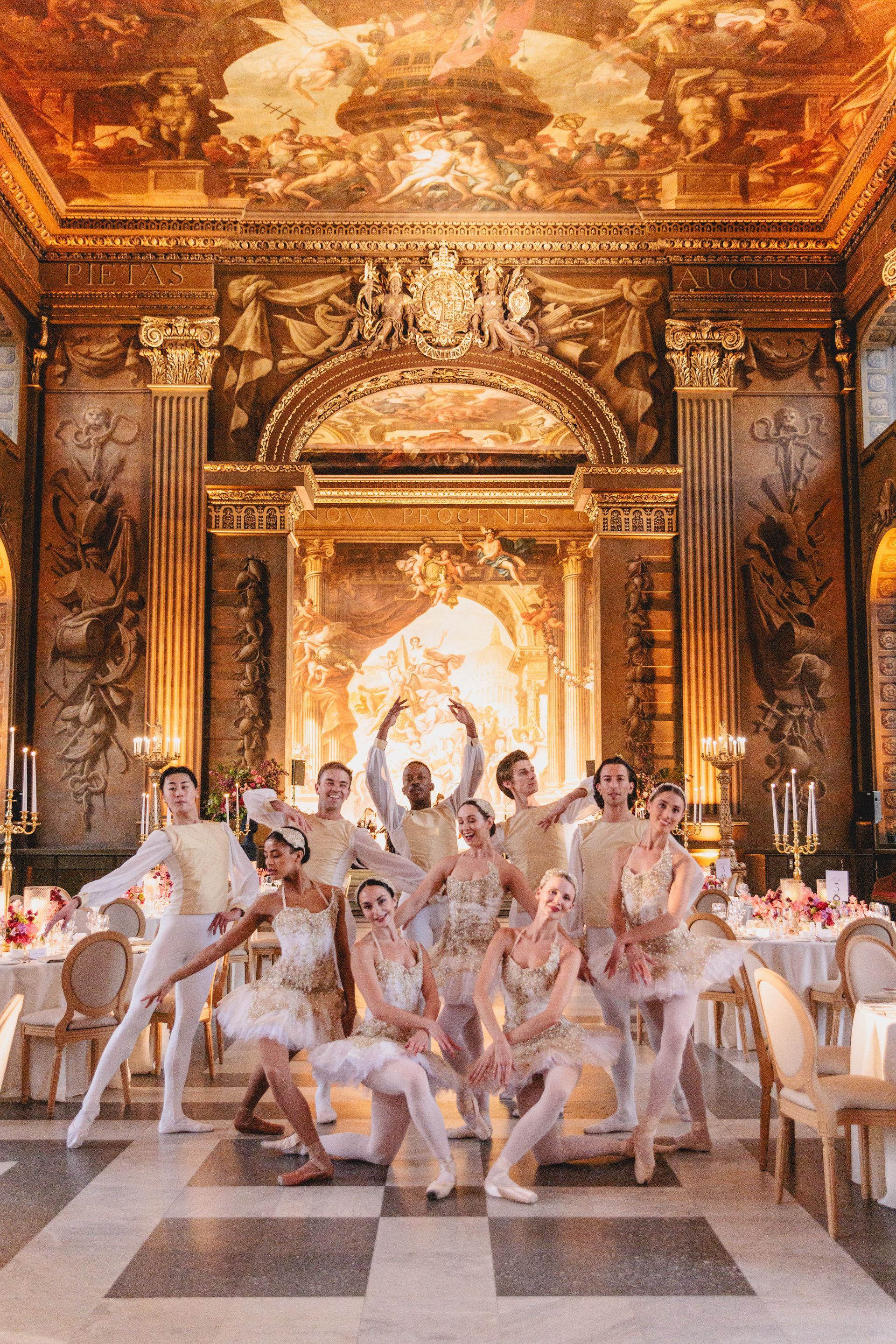 Ballet dancers pose in a gilded room; wearing cream-colored costumes. Tables set with flowers are in the background.