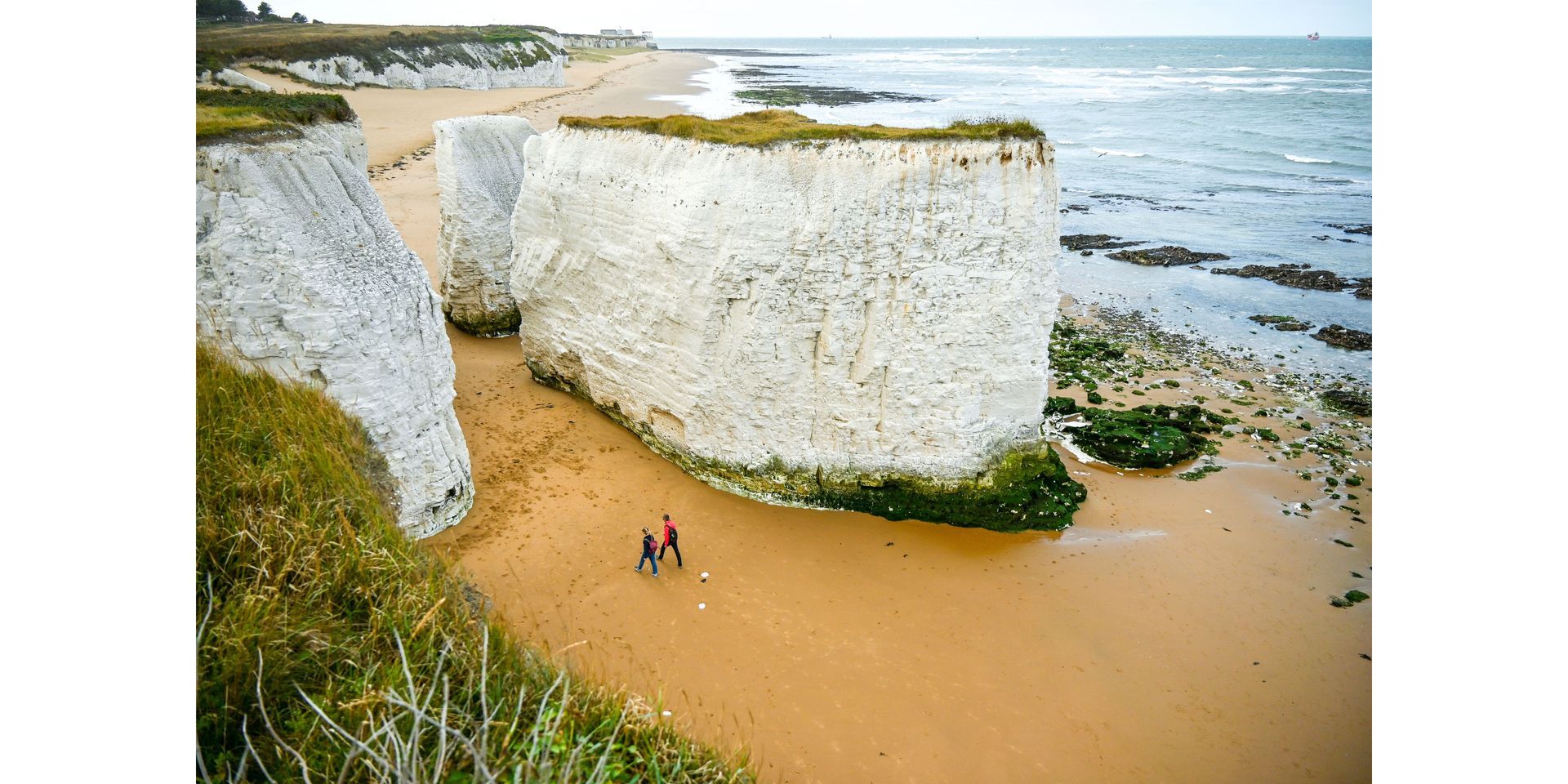 Two people walk on a sandy beach below chalk cliffs with the ocean in the background.