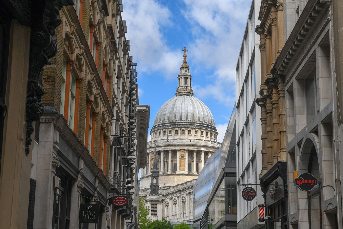 St Paul's Cathedral dome framed by buildings on a London street under a partly cloudy sky.
