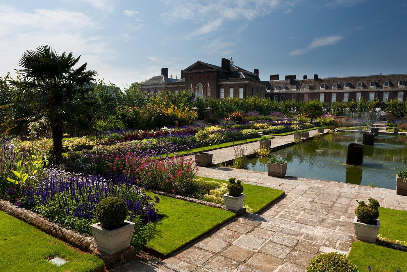 Kensington Palace gardens with flower beds, a pond, and a stone path under a sunny sky.