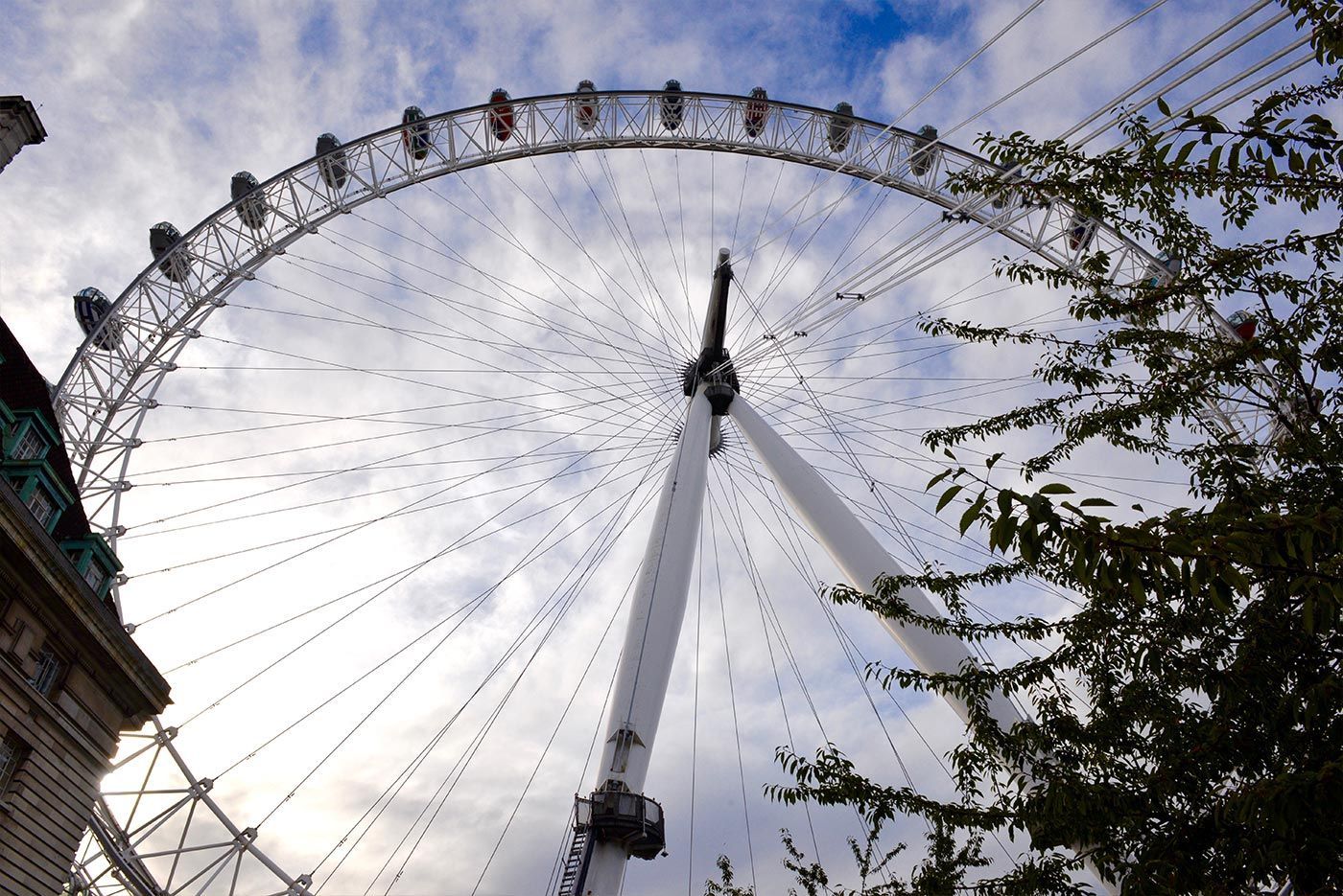 London Eye Ferris wheel, white against cloudy sky, with green foliage to the right and a building on the left.