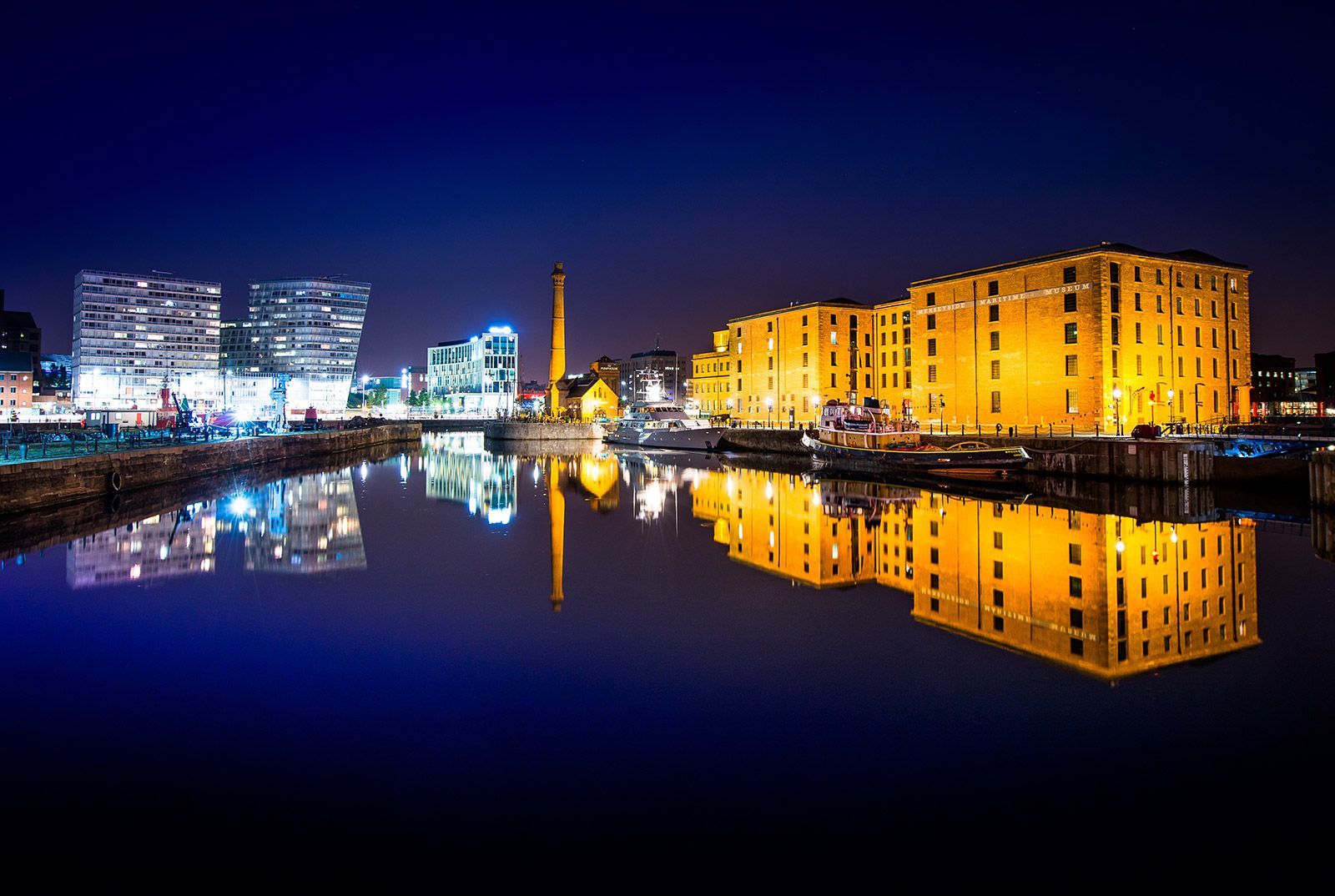 Nighttime cityscape reflects in calm water; buildings glow golden/white against a dark blue sky.