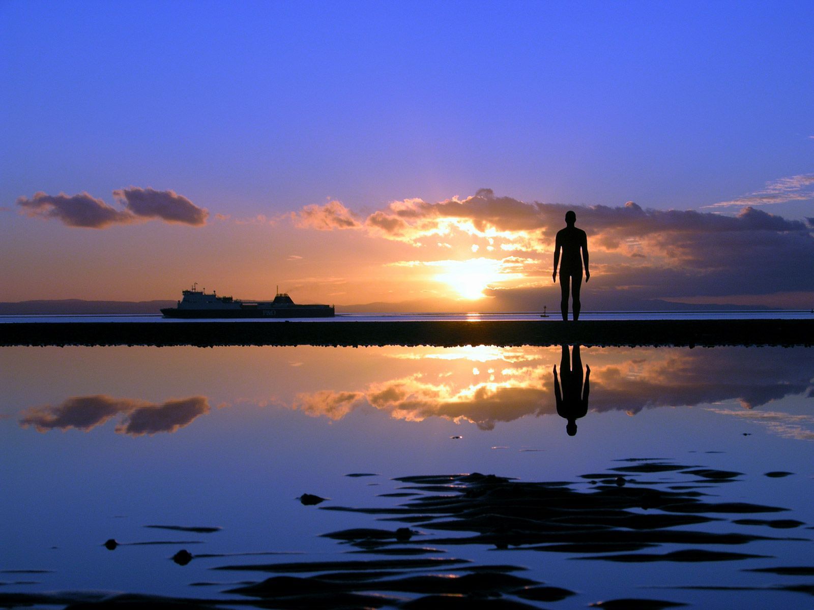 Silhouette of a man on beach, reflecting in water, sunset with ship in distance.