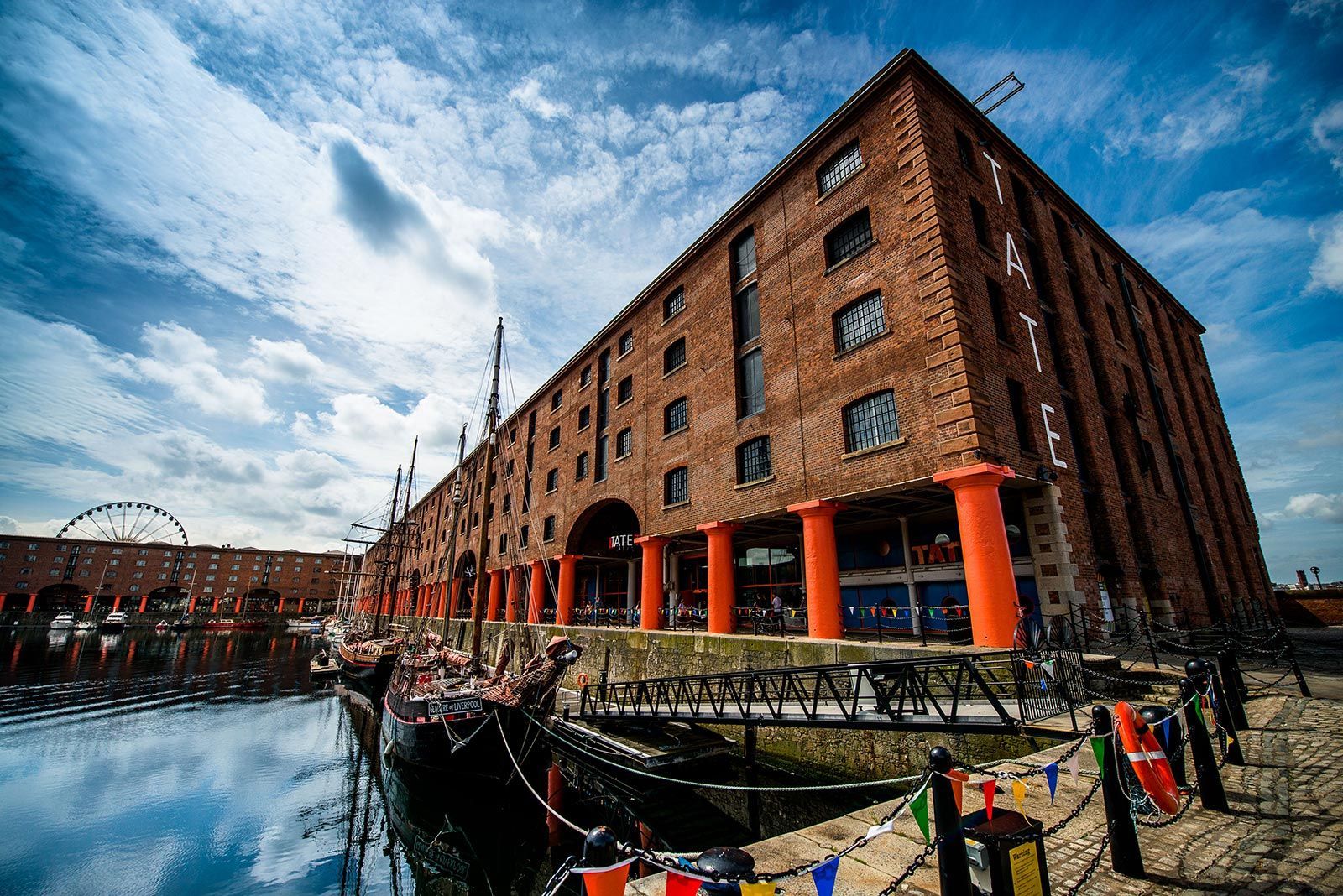 Brick building with orange columns, waterfront, boat, Ferris wheel, cloudy sky.