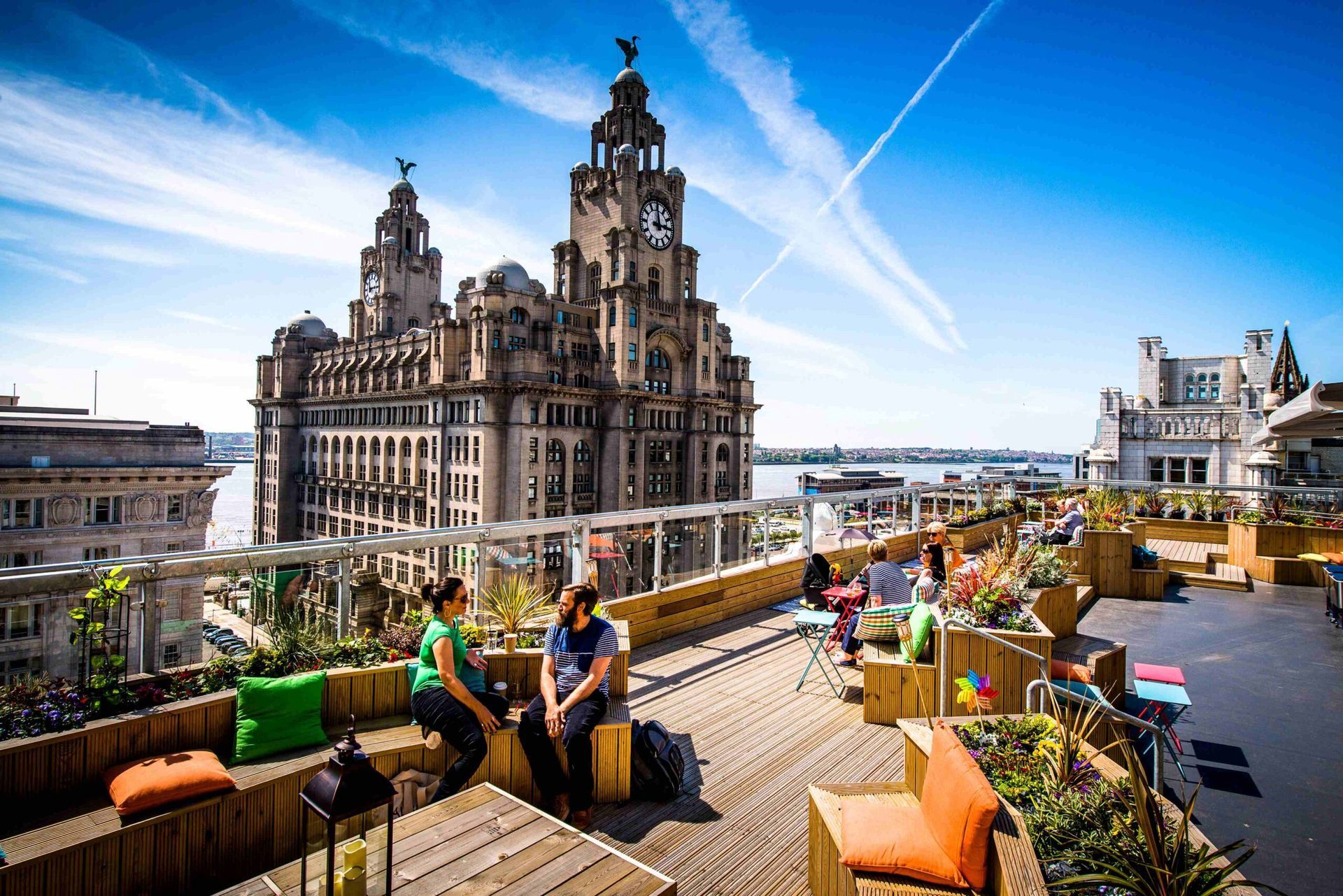 Rooftop bar with people sitting, overlooking Liverpool's Royal Liver Building and waterfront under a blue sky.