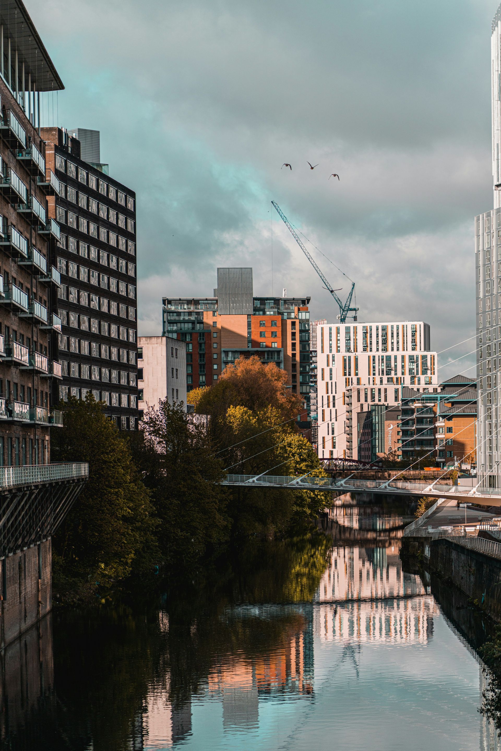 A city skyline with tall buildings reflected in a calm river under a cloudy sky.