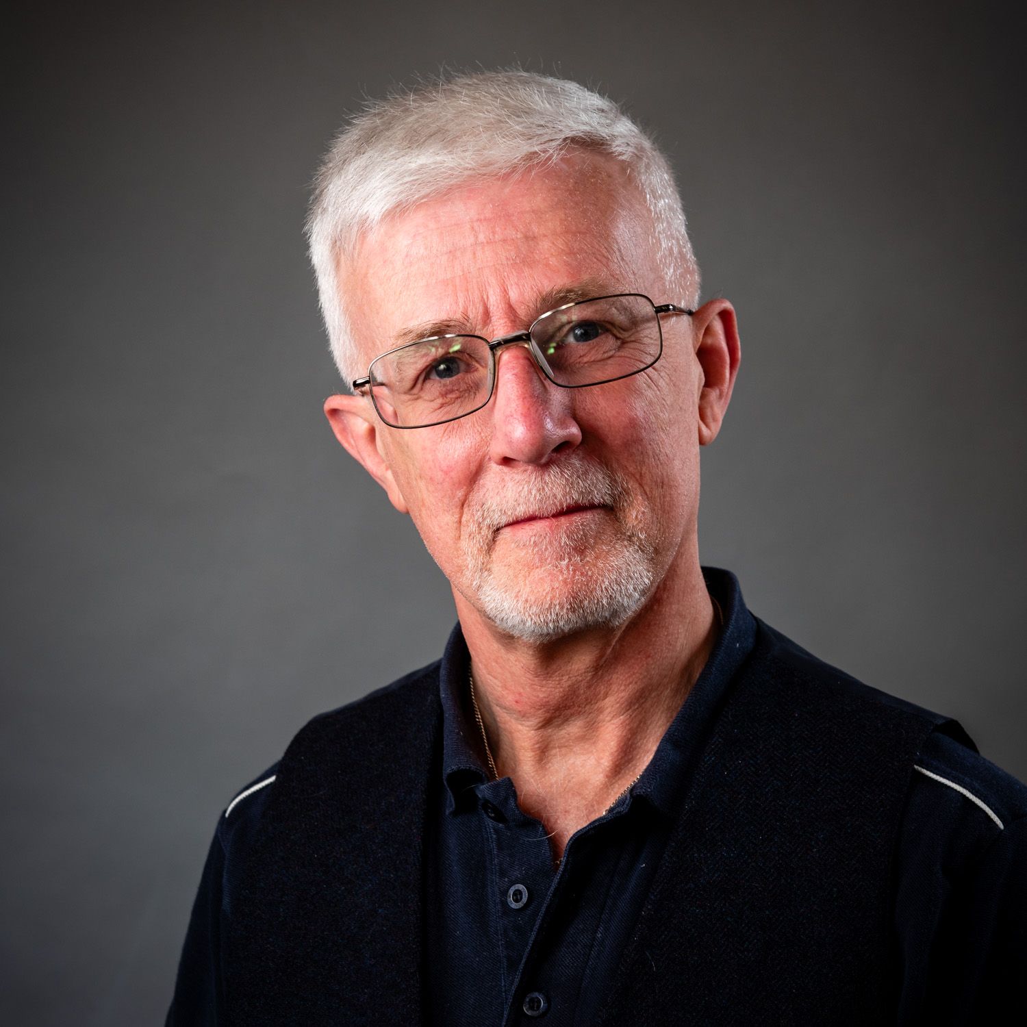 Man with glasses, gray hair, and goatee looking at the viewer. He wears a dark top.