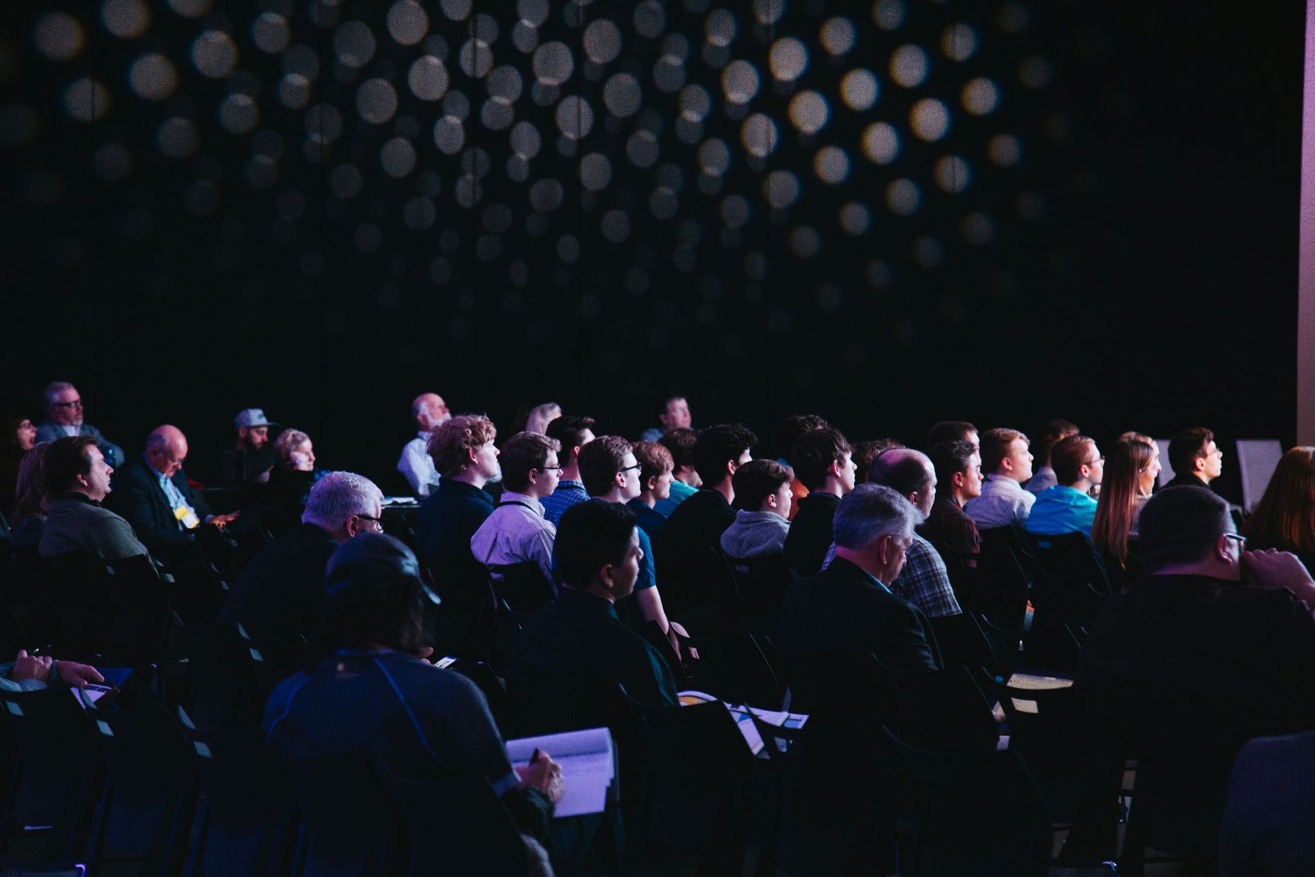 Audience facing a stage, with lights in the background. Many people seated, looking forward.