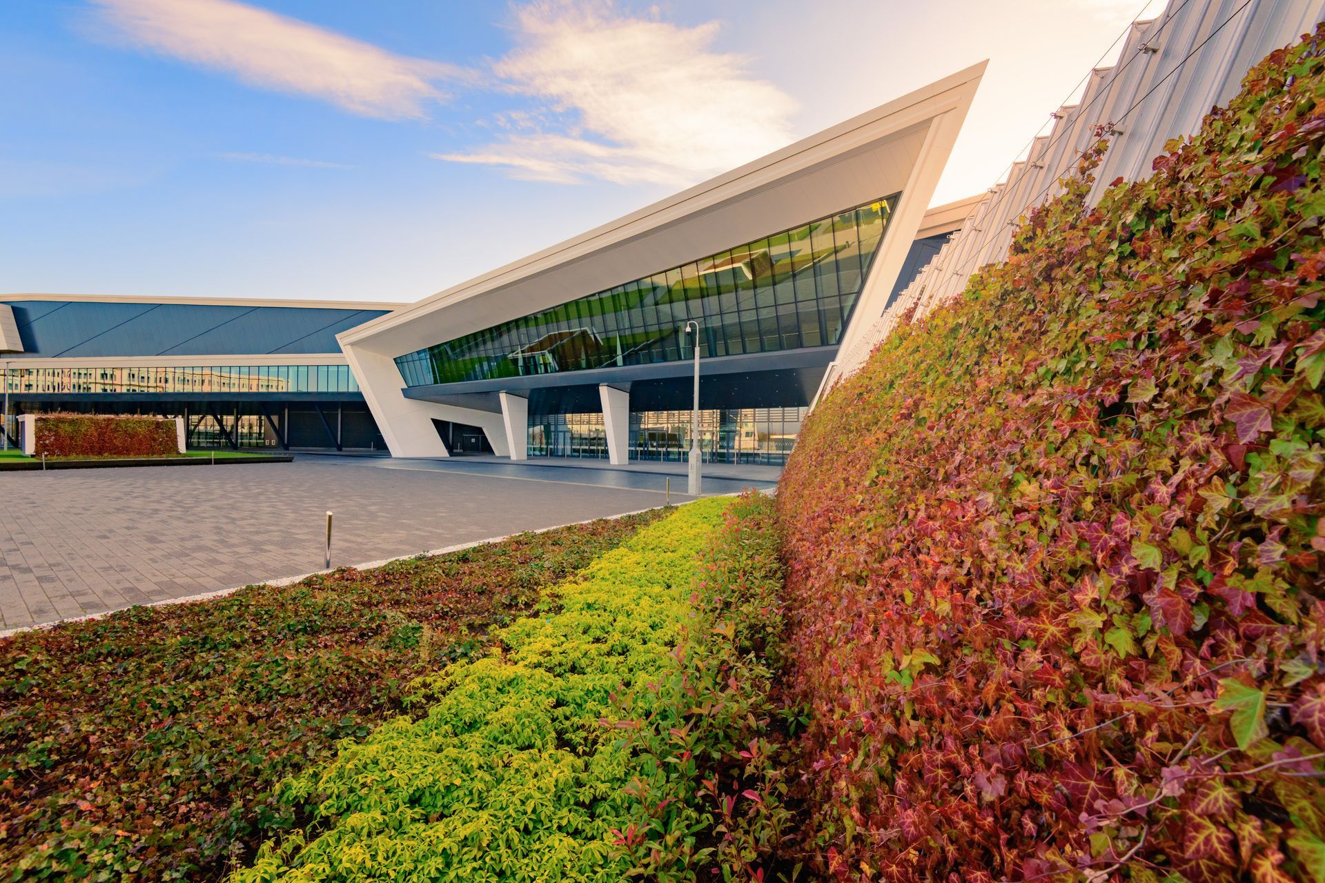 Modern building with angled white roof, glass windows, and green/red foliage. Sunny day, blue sky.
