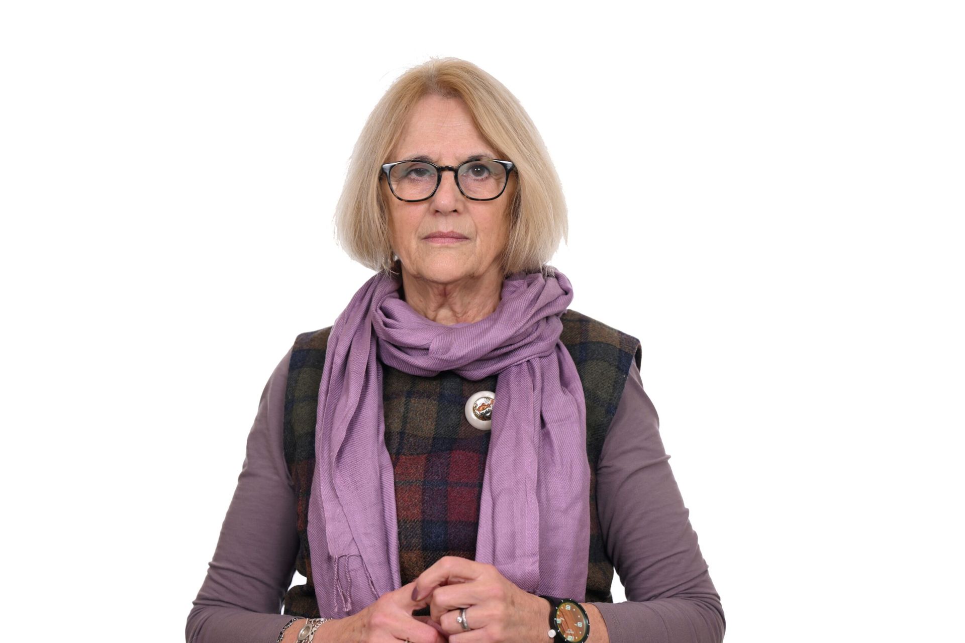 Woman wearing glasses, purple scarf, and plaid vest, looking forward with hands clasped.