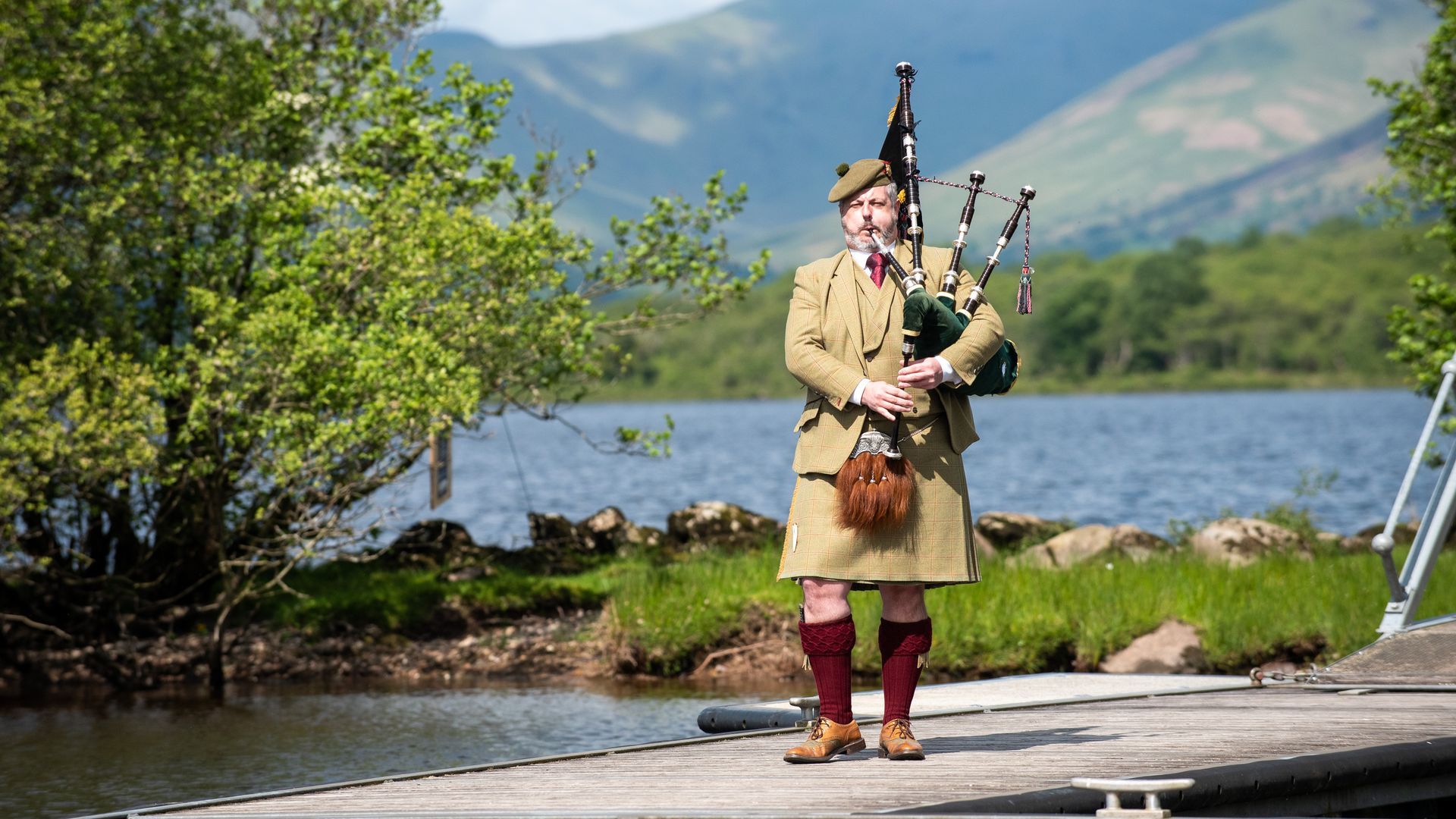 Man in kilt playing bagpipes on a dock, lake and mountains in the background.