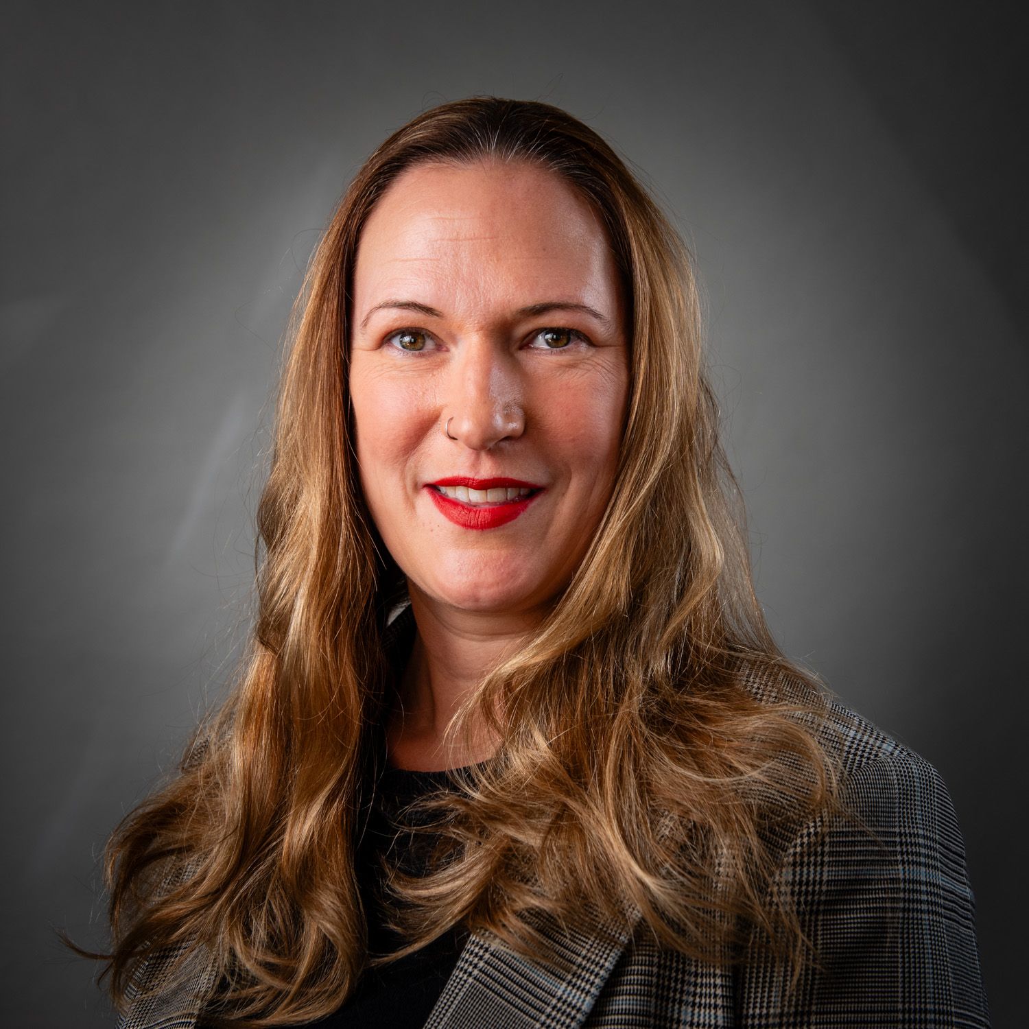 Woman with long brown hair, wearing a patterned blazer and red lipstick, smiles against a gray backdrop.