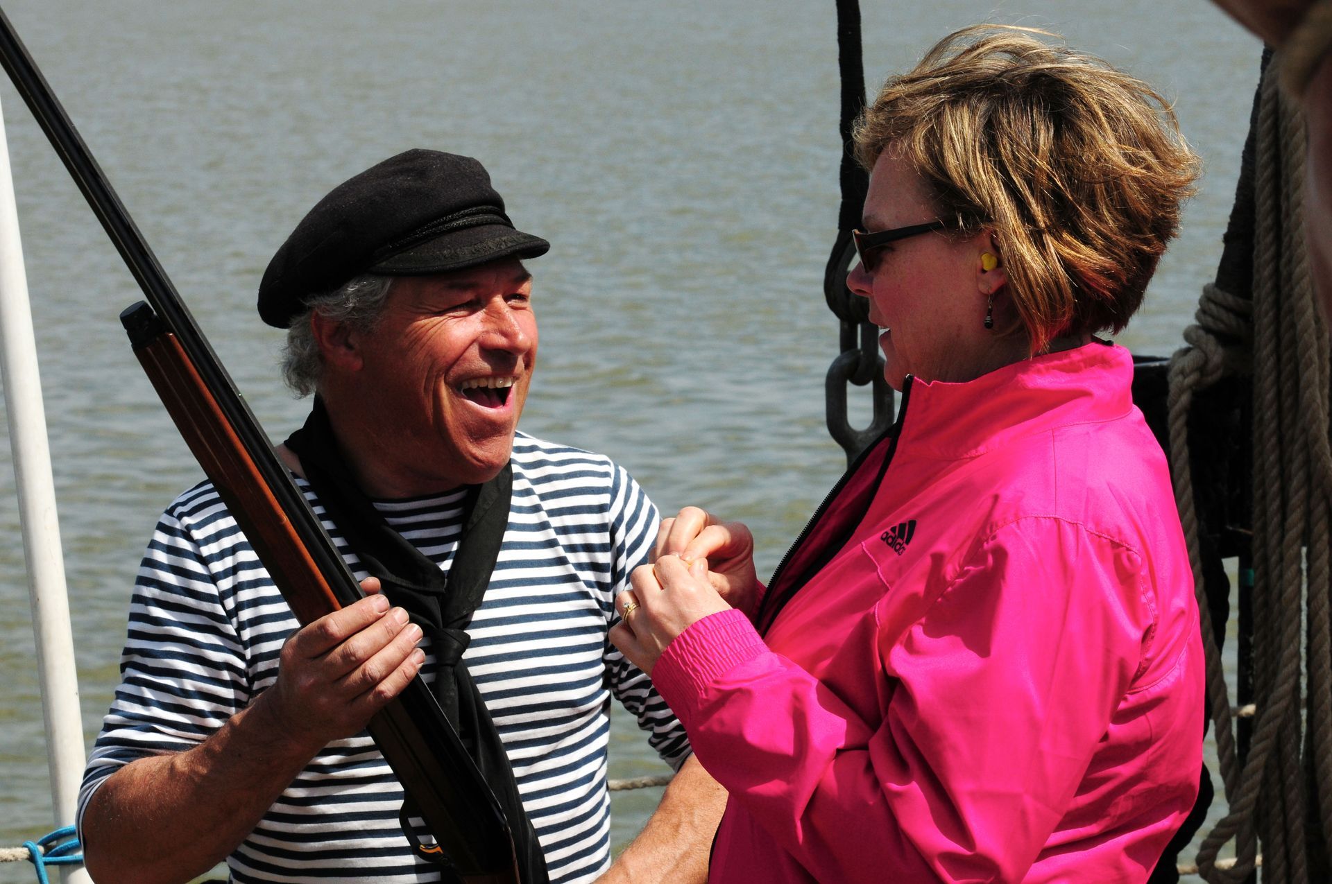 Man in sailor hat laughs while holding a rifle, talking to a woman in pink jacket on a boat.