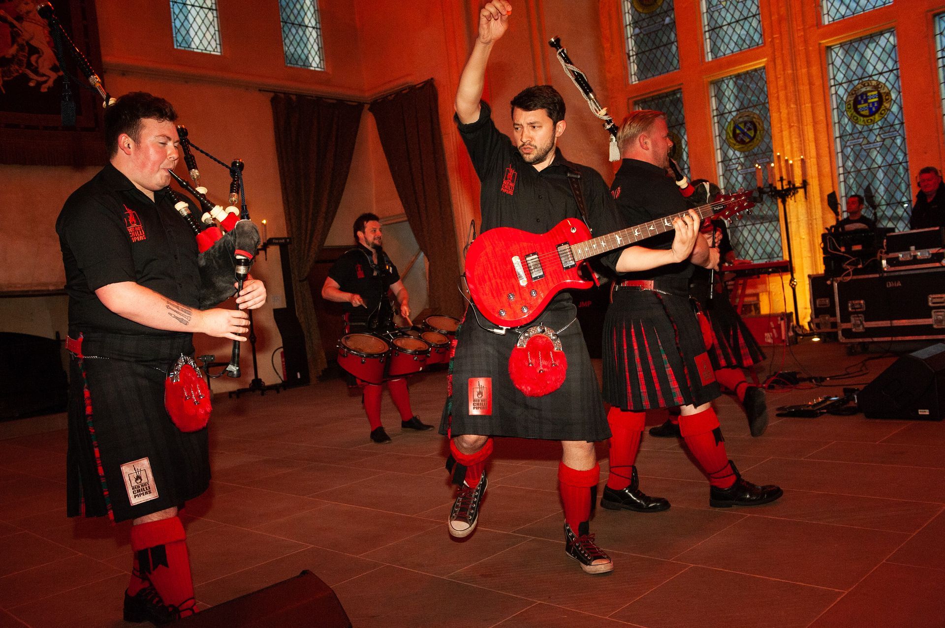Band in kilts playing music; bagpipes, guitar, drums. Indoor performance with red and black.