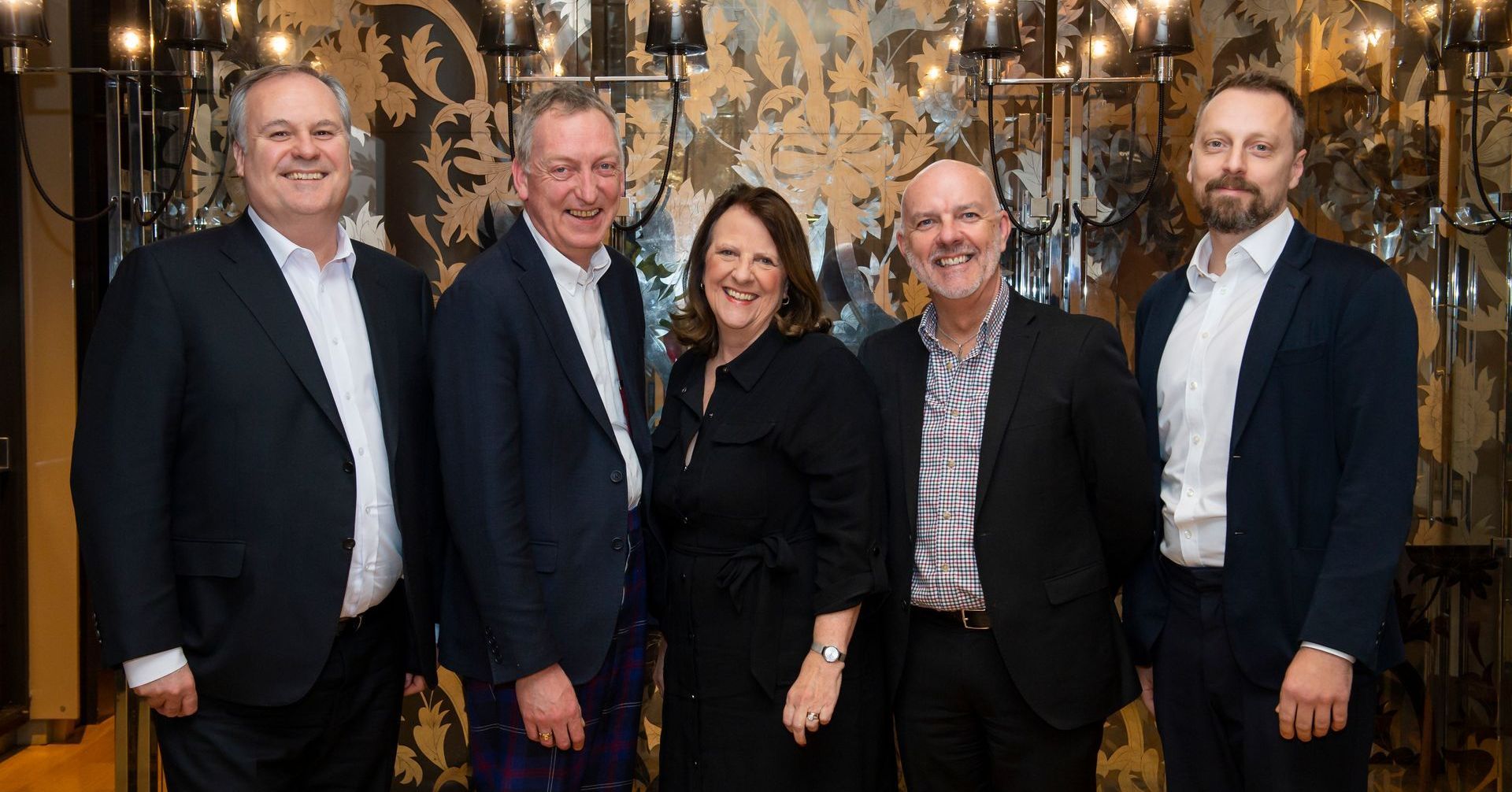Five people smiling, standing, indoors in front of ornate wall. Men in suits, woman in black dress.