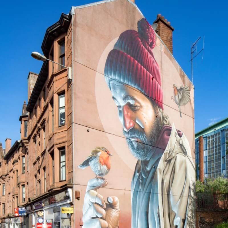 Mural of a man with red beanie interacting with robin on a building wall in Glasgow.