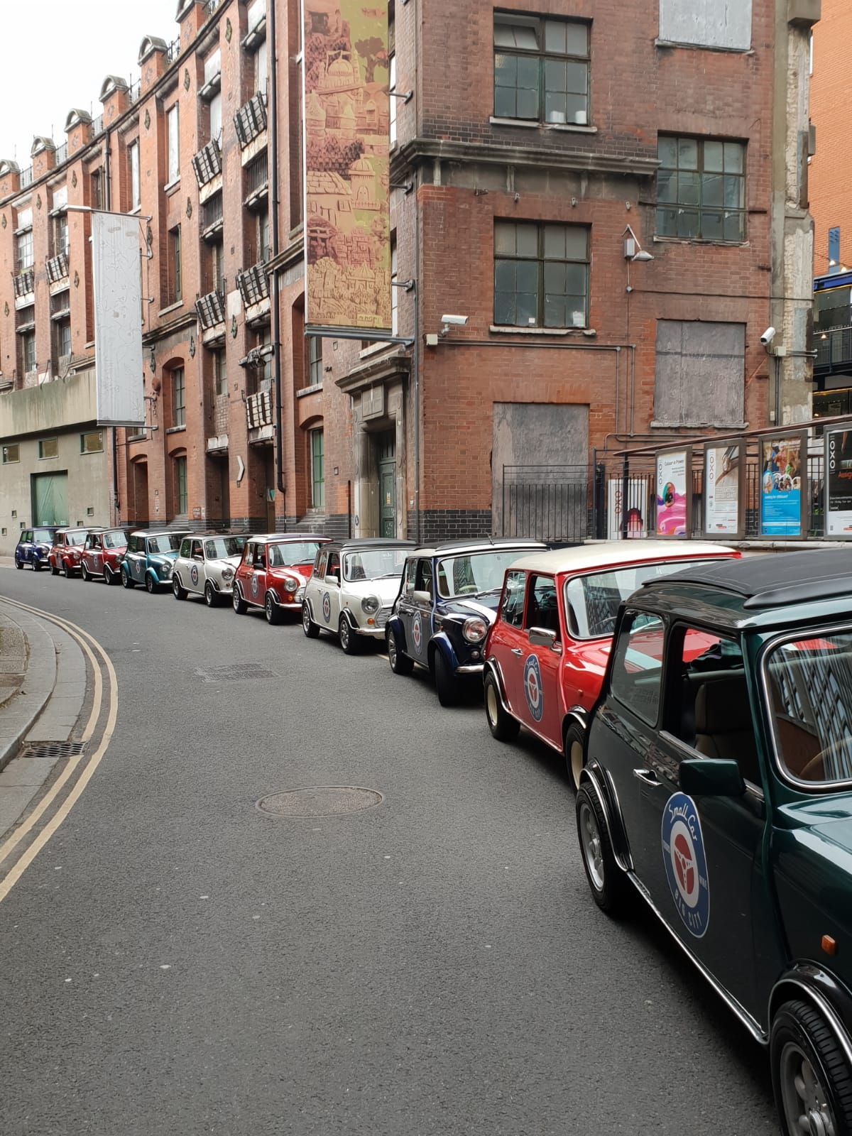 Row of classic Mini Coopers parked on a narrow city street, surrounded by brick buildings.